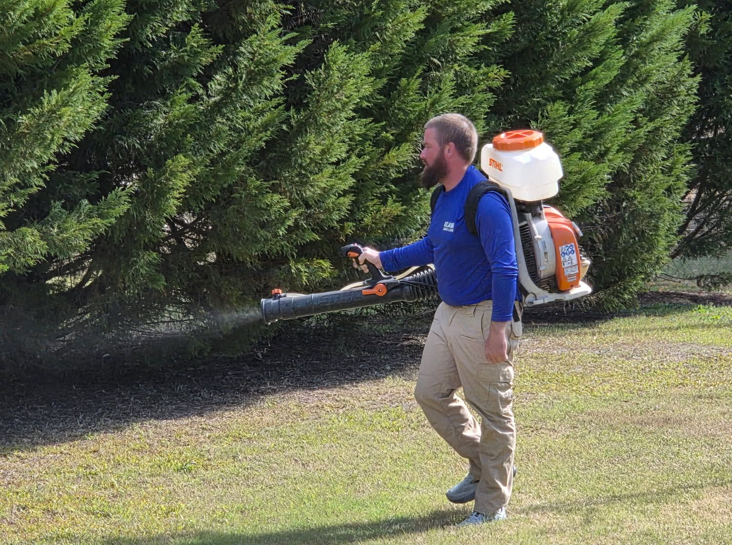 A professional worker wearing a blue uniform and khaki pants uses a backpack sprayer to treat a hedge along a lawn.