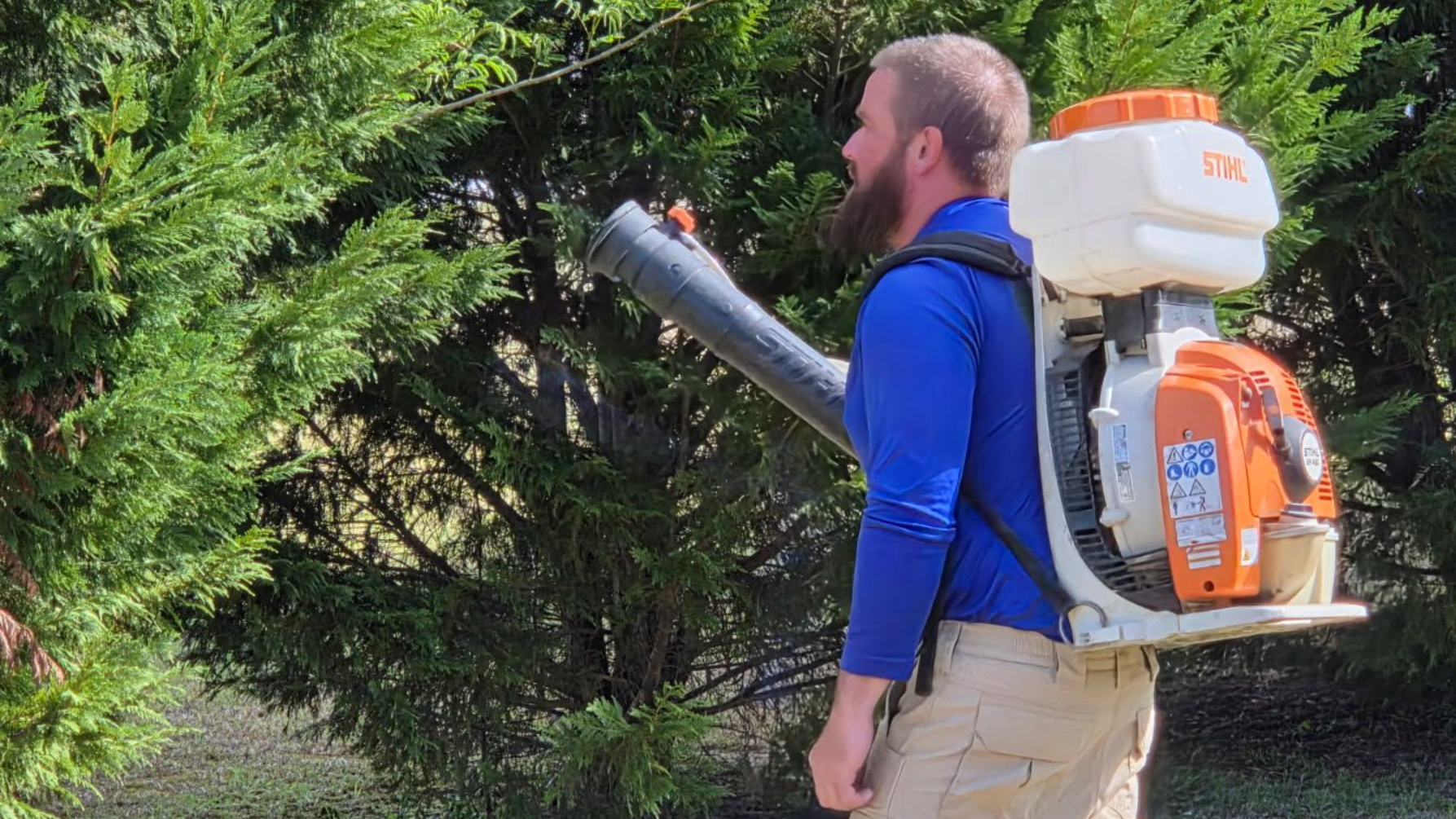 A person wearing a blue shirt and khaki pants carries a backpack-style leaf blower while working near green bushes.