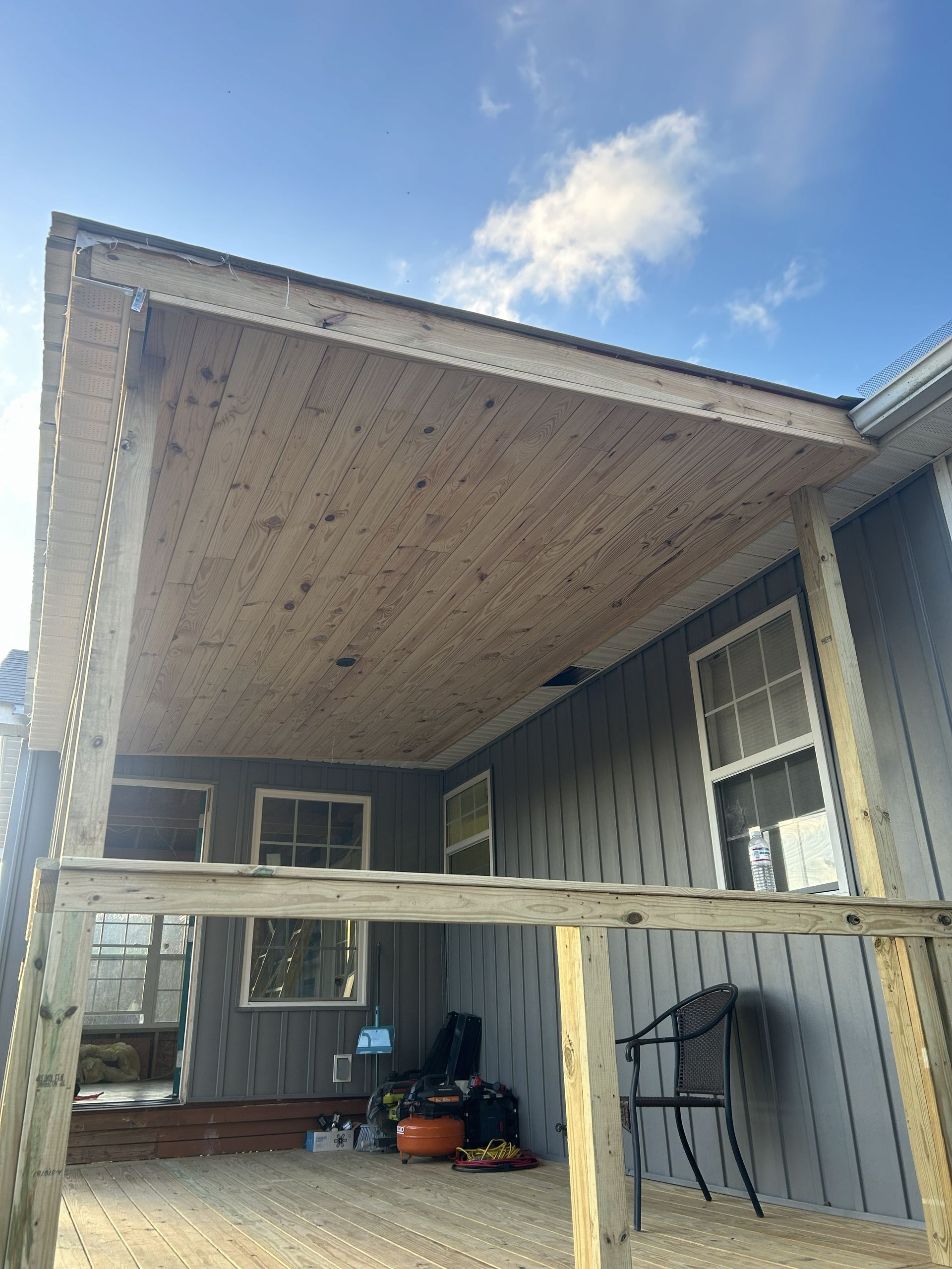 Covered porch with exposed ceiling, gray siding, and storage items under a partly cloudy sky