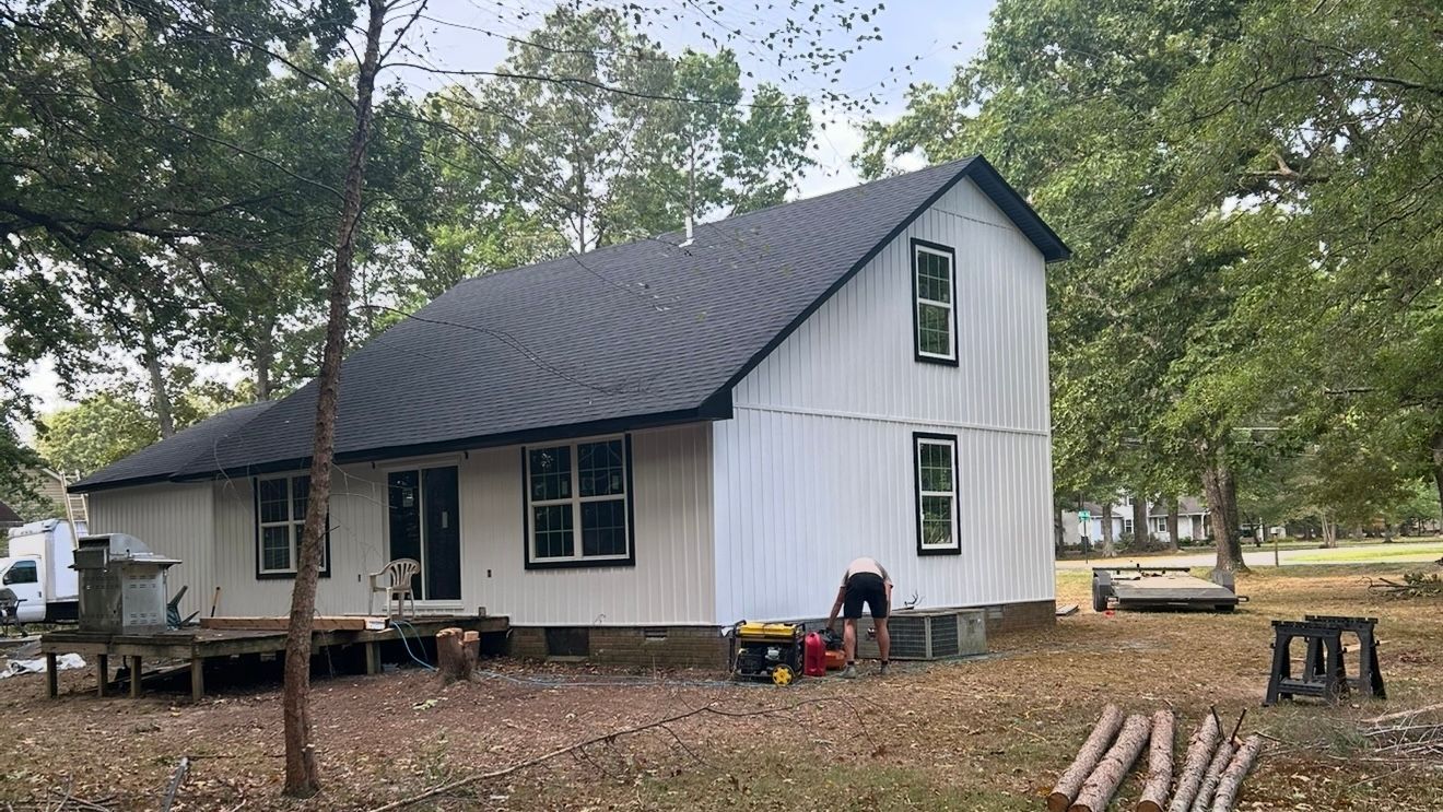 White house with dark metal roof in a wooded yard, with trees and a porch under construction.
