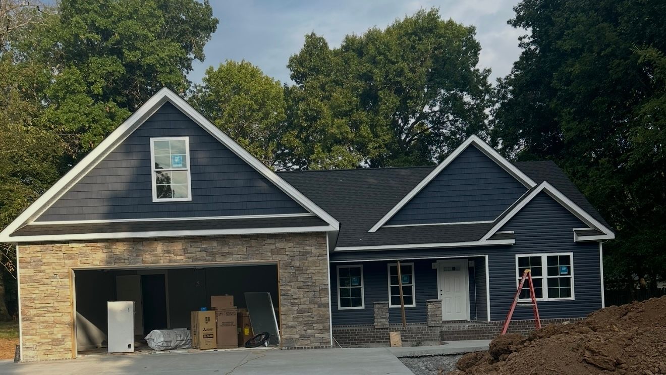 New gray house with stone garage and front porch under construction, trees in the background.