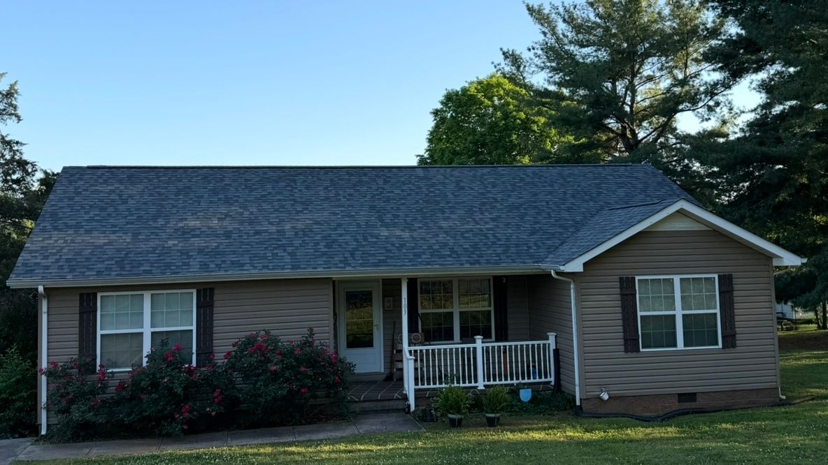 Single-story house with gray siding, dark roof, white porch railing, and front shrubs under trees