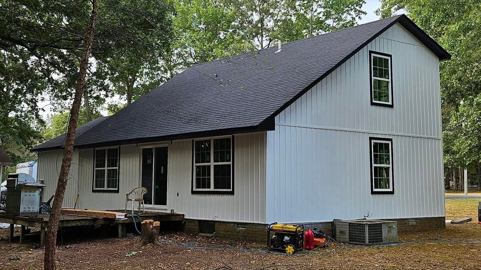 White farmhouse with dark roof and porch in a wooded yard, with trees and scattered outdoor items.