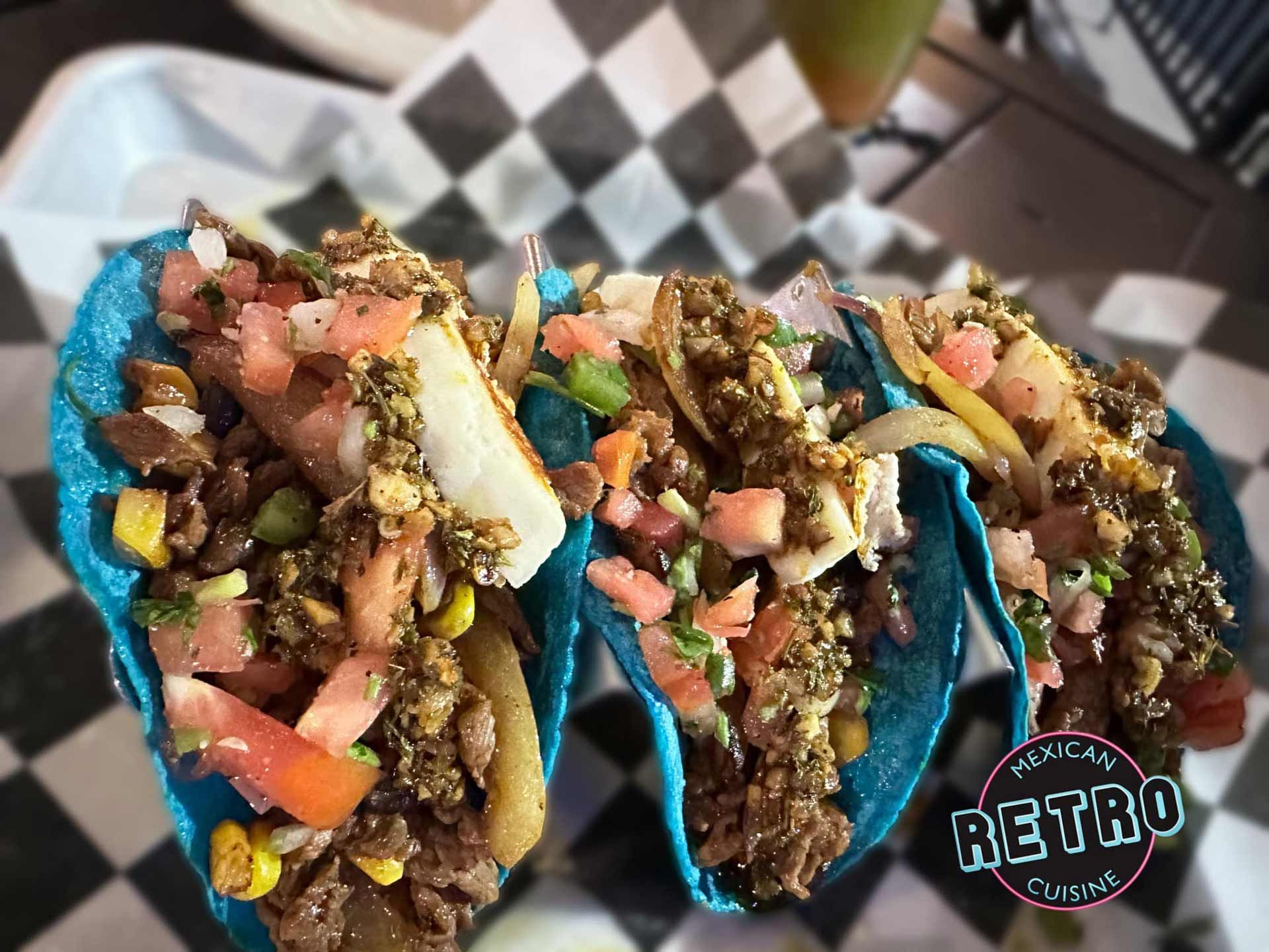 A row of blue tacos with meat and vegetables on a checkered table cloth