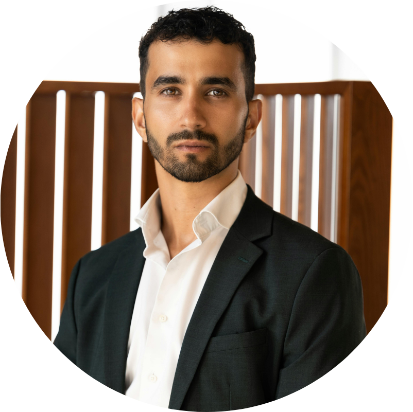 Man in a suit, staring directly at the camera. Light skin, short beard. Set against a wooden backdrop.