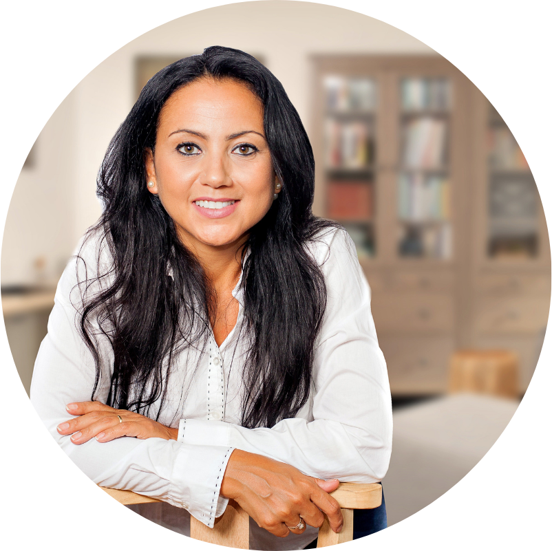 Woman with dark hair smiles at the camera while leaning on a chair in a well-lit room.