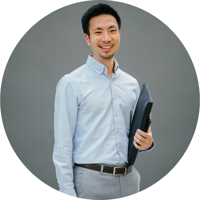 Smiling Asian man in light blue shirt, holding a dark folder, against a gray backdrop.