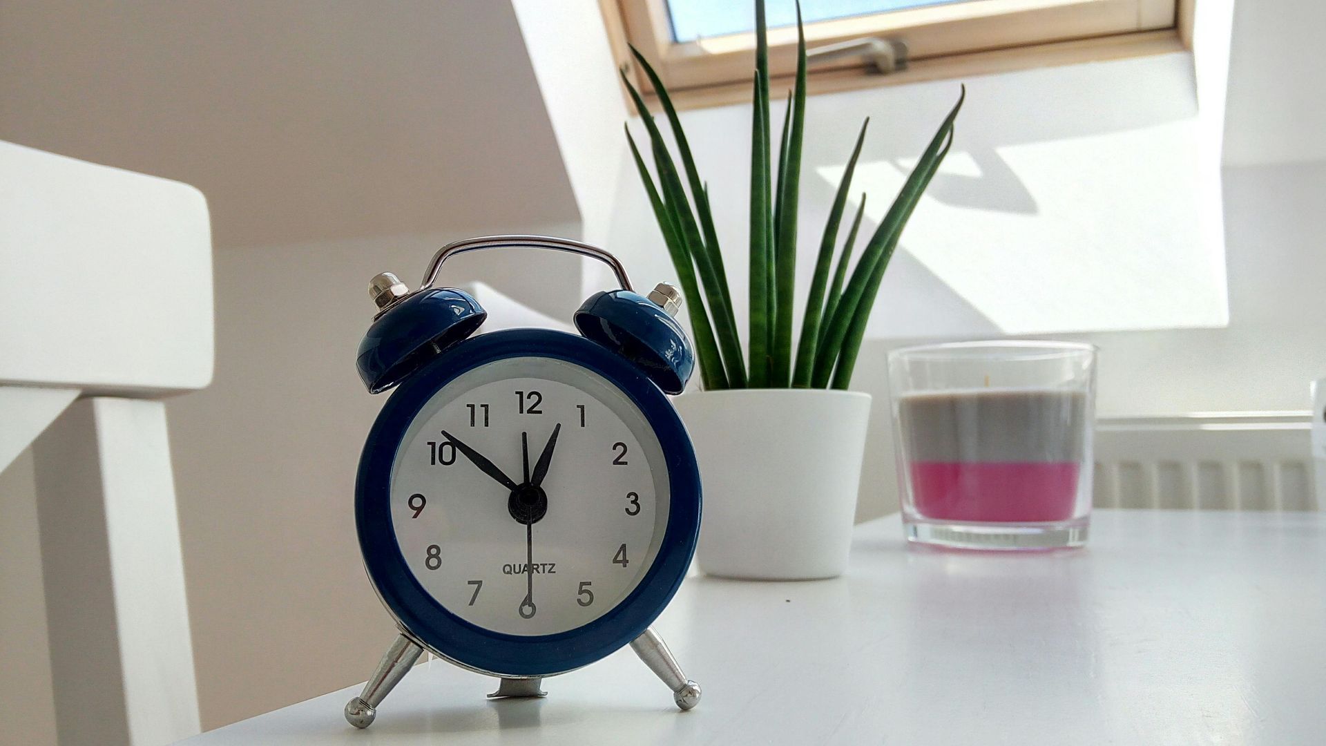 A blue alarm clock is sitting on a table next to a plant and a candle.