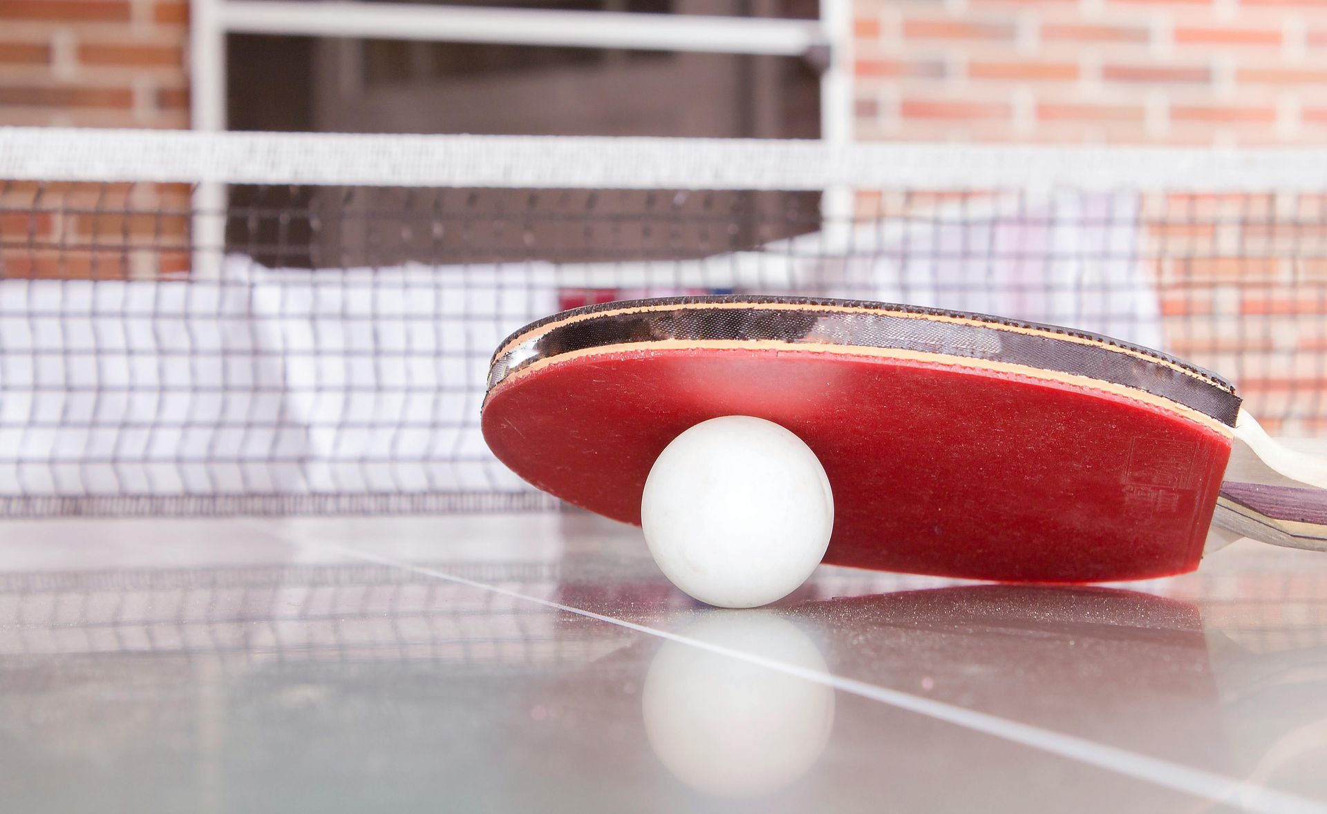 Ping pong paddle and ball on a table, with net and brick wall background.
