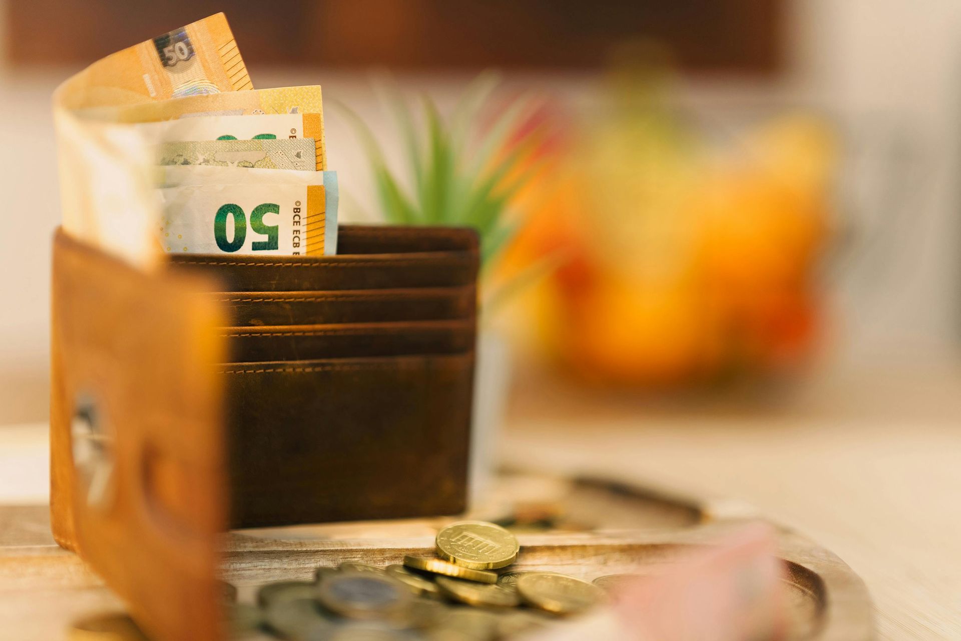 Brown wallet with Euro banknotes and coins on a wooden surface; blurred background with fruit and greenery.