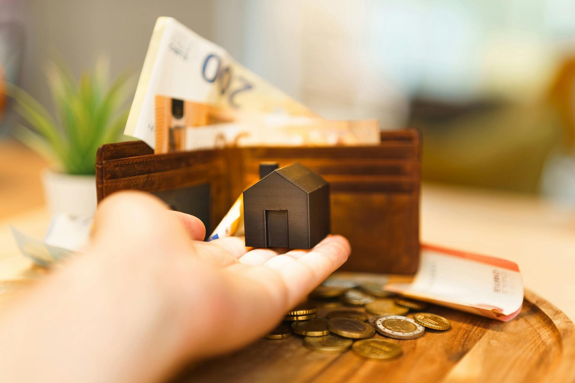 A hand holds a small black house model in front of an open wallet filled with Euro banknotes and loose coins on a table.