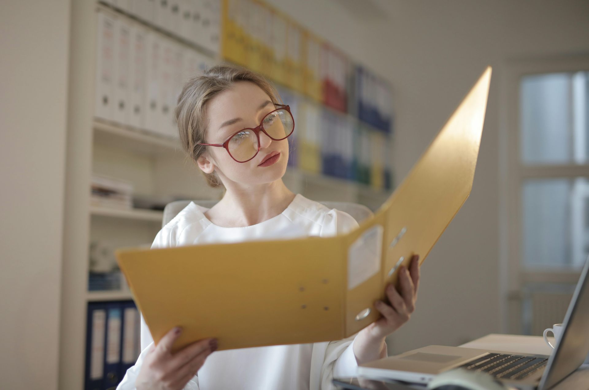 Woman in glasses reading a yellow folder at a desk in a bright office