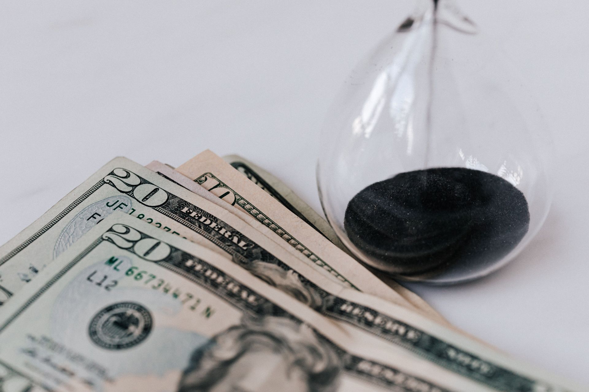 Dollar bills next to a small hourglass with black sand on a white surface
