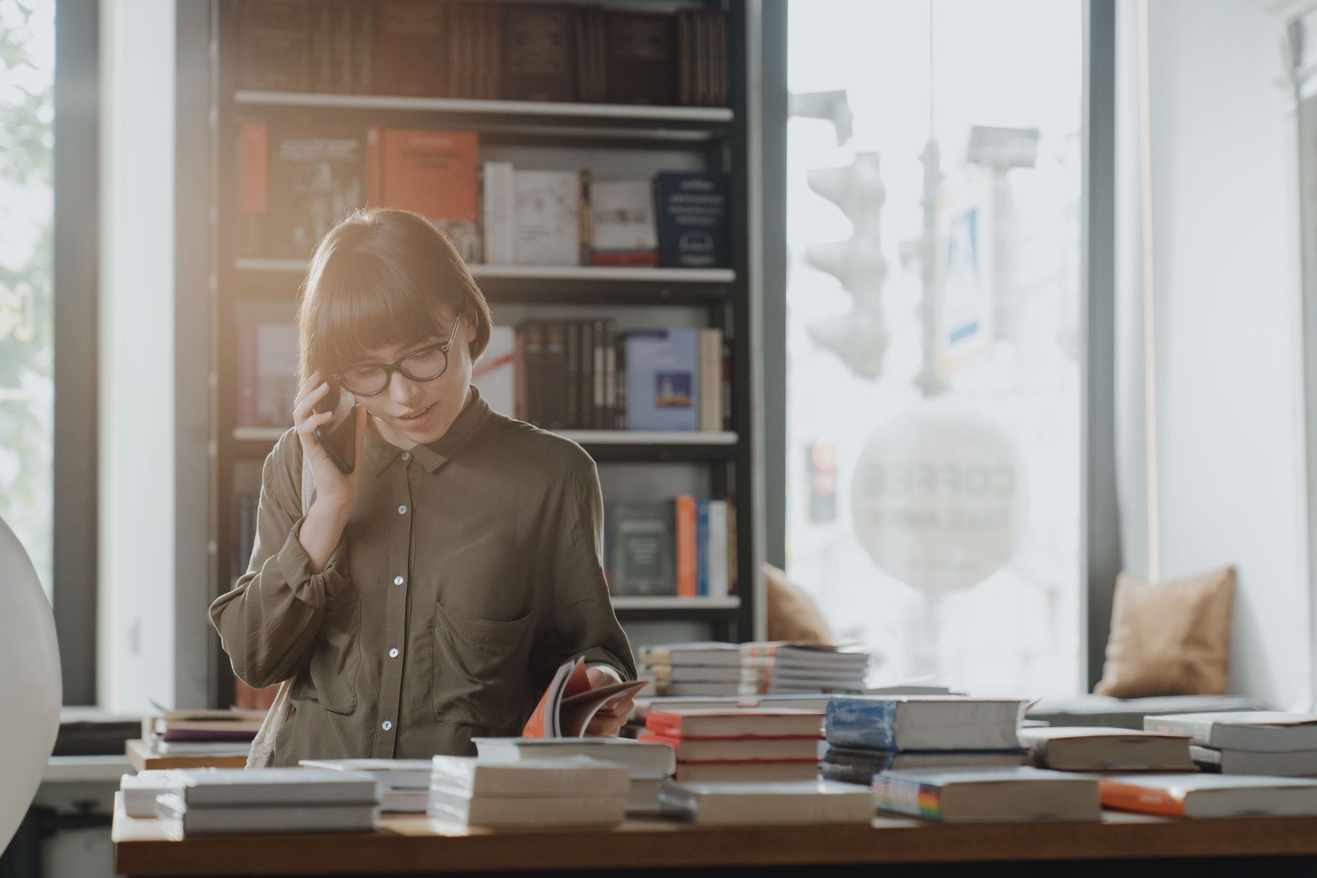 Person talking on the phone in a bright office, standing behind a desk with stacks of books and papers.