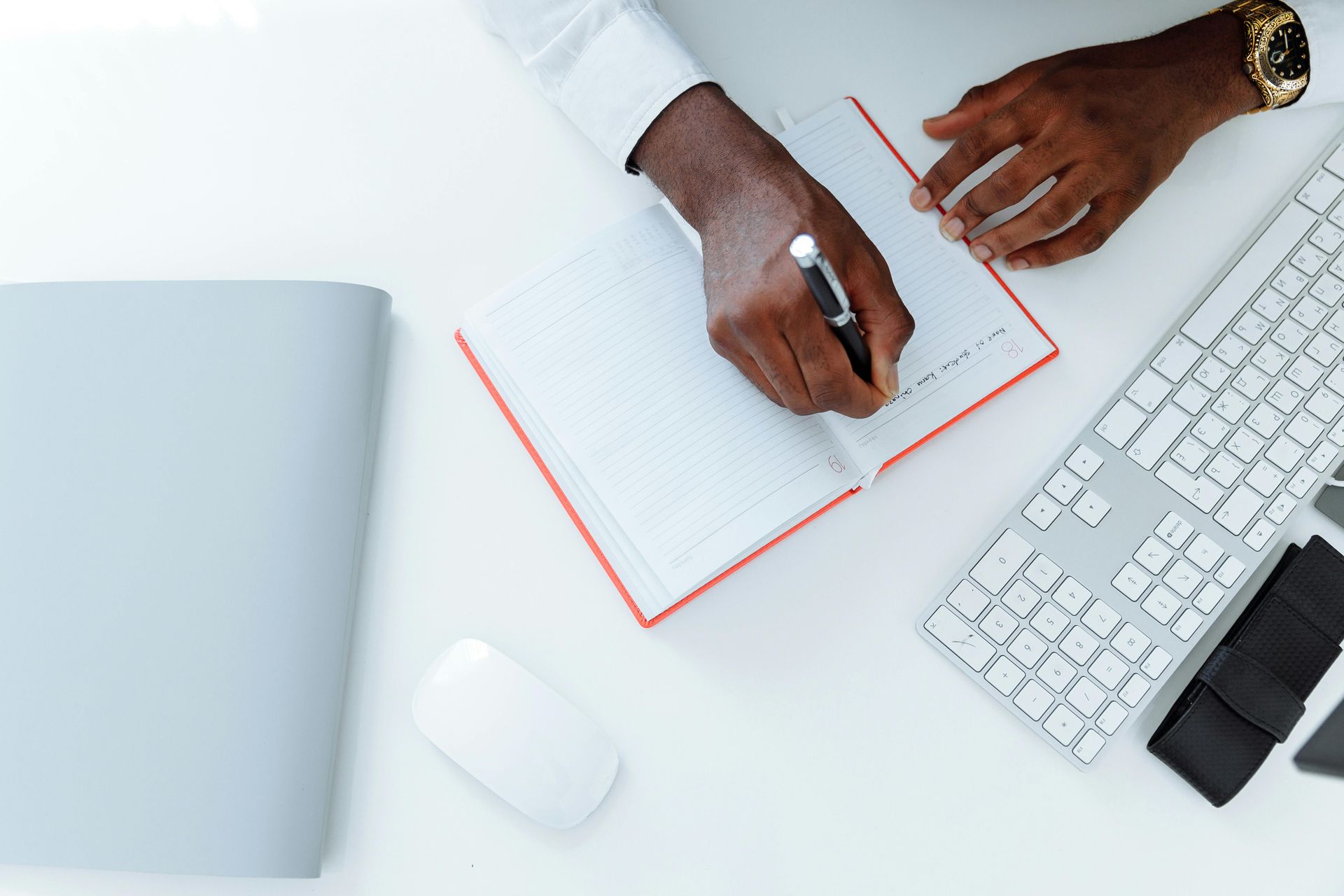Hands writing in a notebook next to a keyboard, mouse, and folder on a white surface.