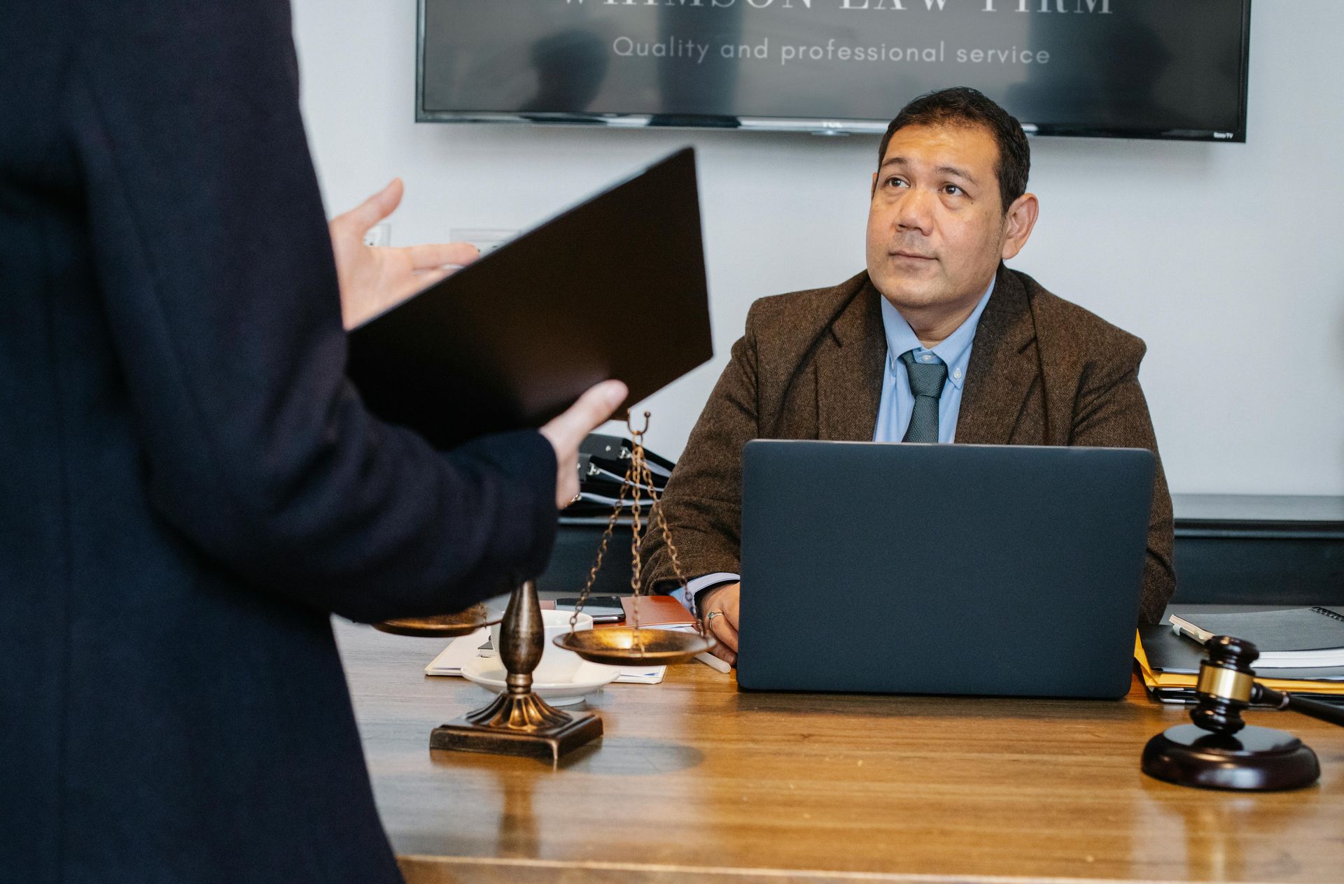 Lawyer in a brown suit listens intently at a desk; another person gestures, holding a folder.