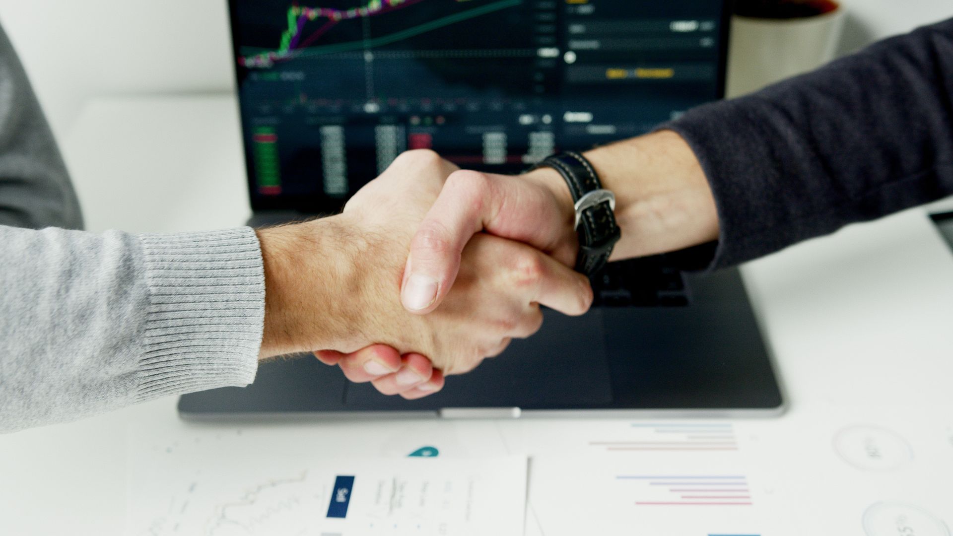 Two people shaking hands over a laptop with charts on screen, in a business setting