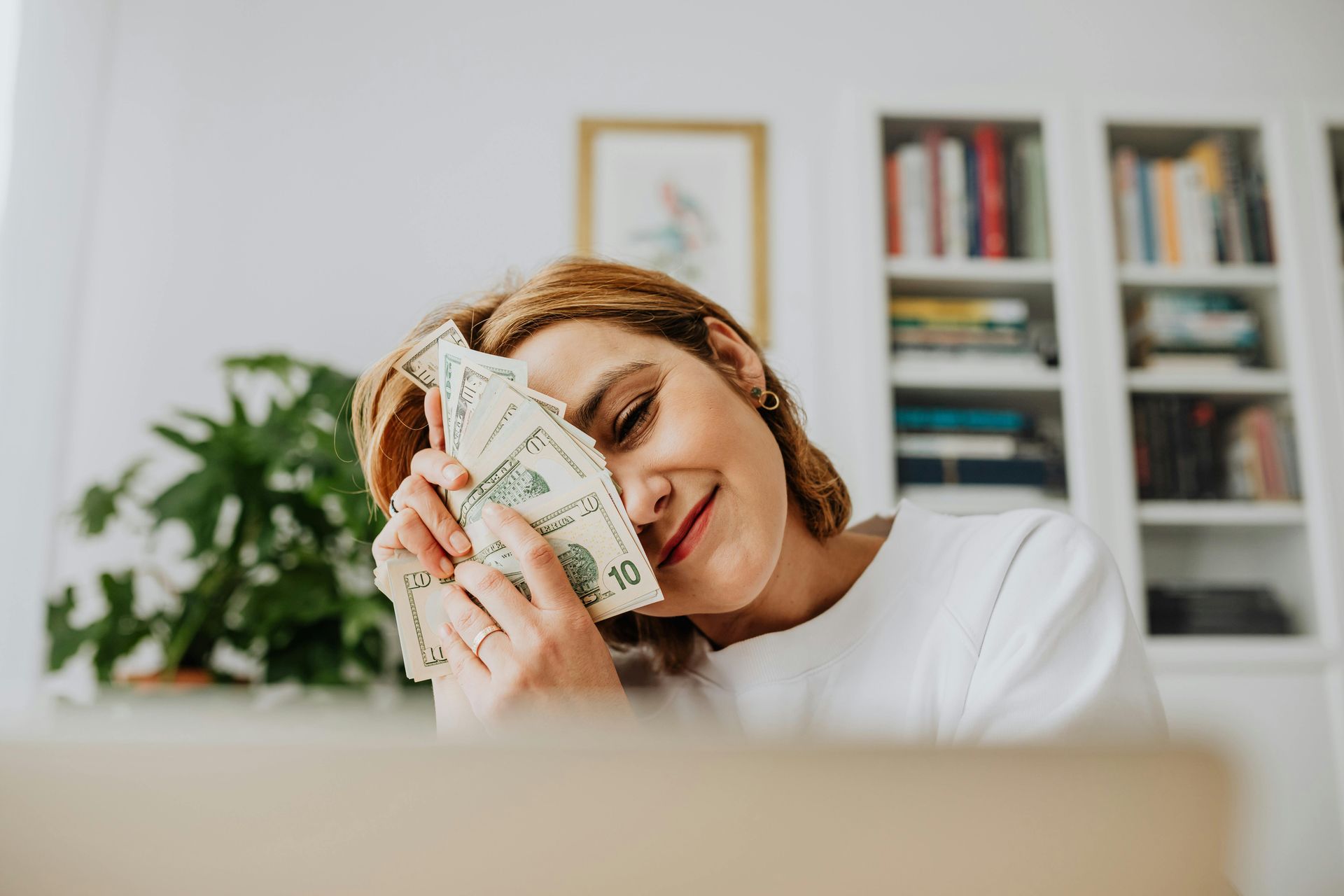 Woman smiling, holding cash, near a laptop and bookcase.