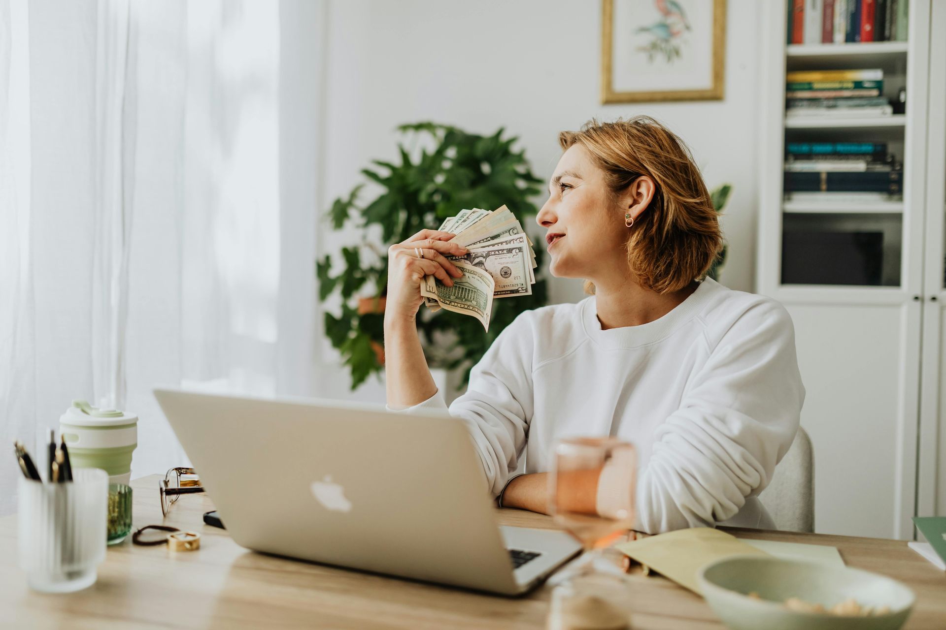 Woman holding cash, looking thoughtful, sitting at a desk with a laptop, indoor setting.