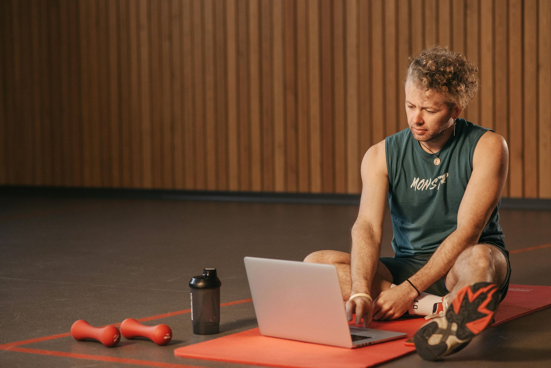 Man in athletic wear using laptop on a yoga mat with weights and water bottle in a wood-paneled room.