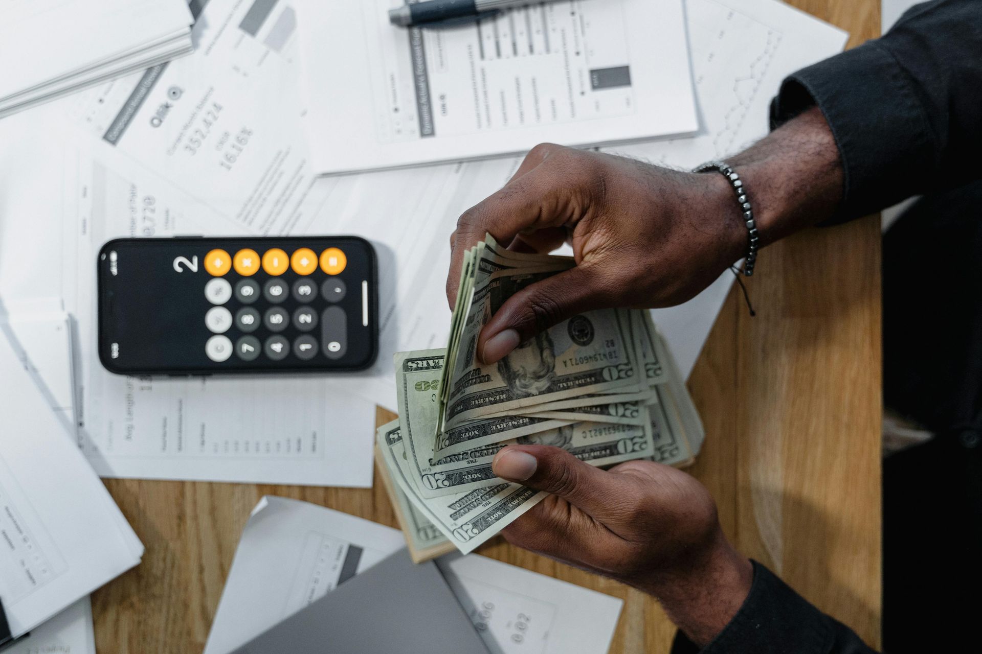 Person counting cash with a calculator and paperwork on a wooden desk.