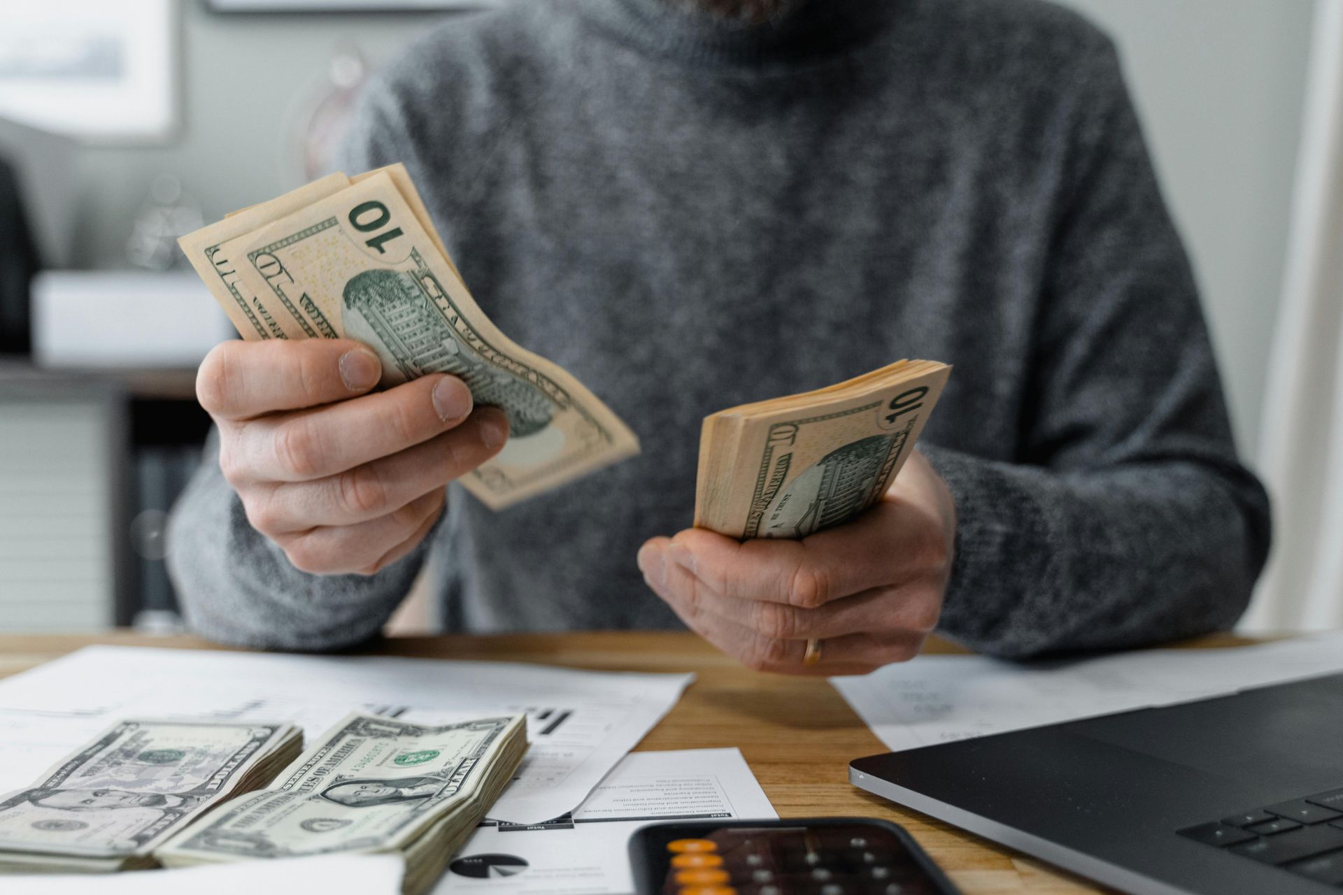 Person counting cash at a table with stacks of money, calculator, and laptop nearby