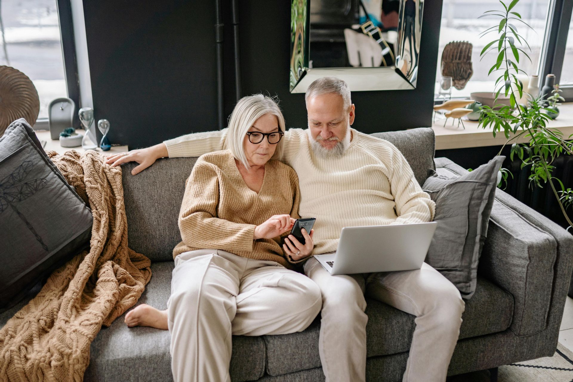 Elderly couple on a gray couch looking at phone and laptop; living room setting.