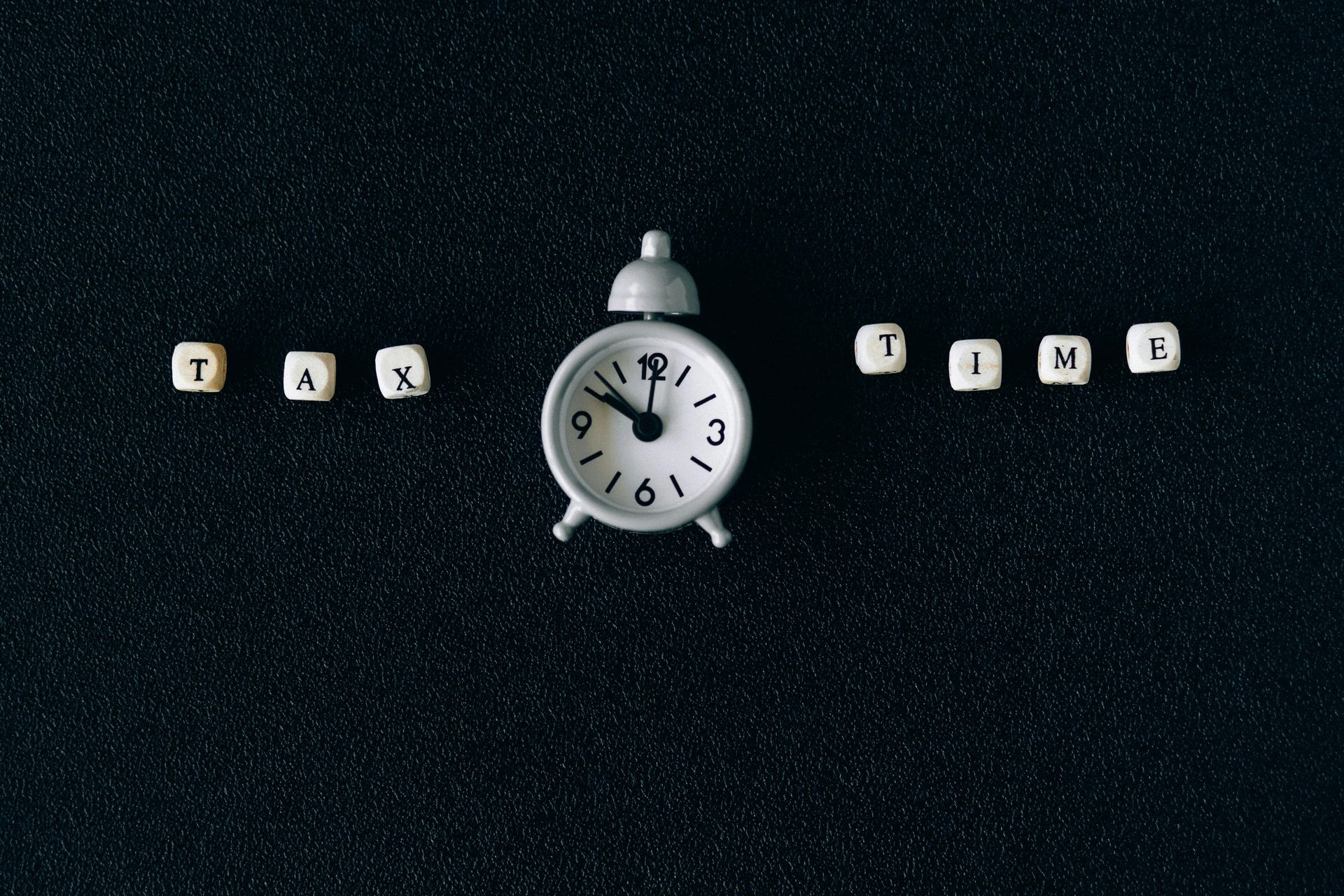 White alarm clock between letter tiles spelling “TAX” and “TIME” on a dark background