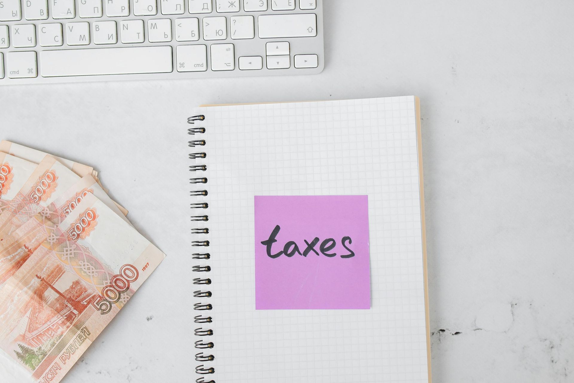 Notebook with a pink sticky note labeled “taxes” beside euro banknotes and a keyboard on a desk