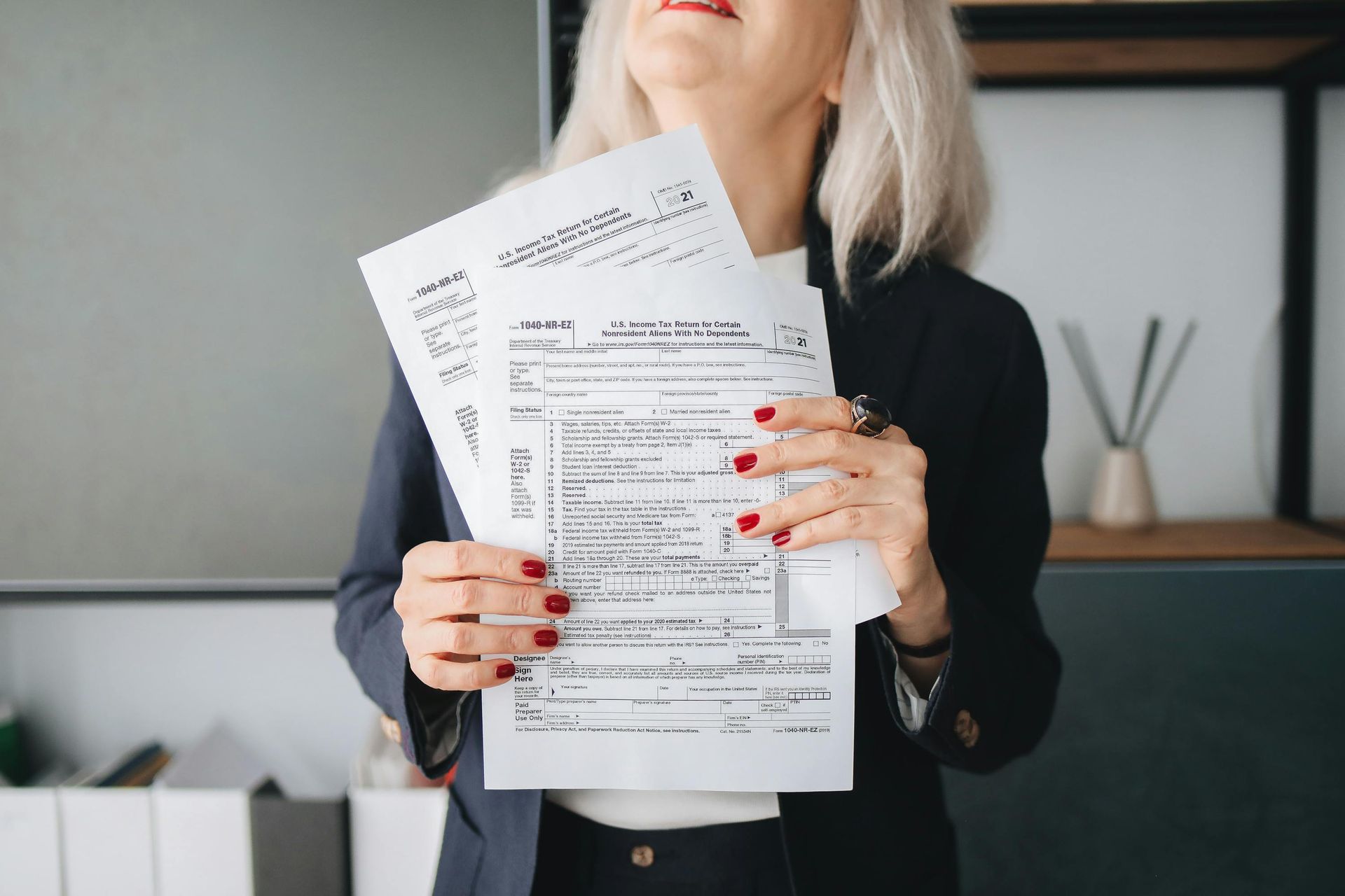 Woman in blazer holding paperwork, looking upwards with a slight smile. Red nail polish, office setting.