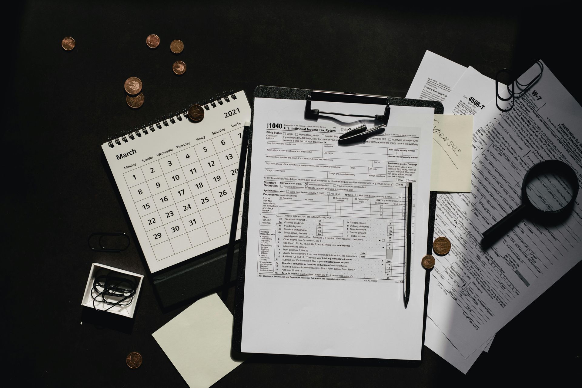 Desk with calendar, clipboard with notes, magnifying glass, papers, coins, and small box.