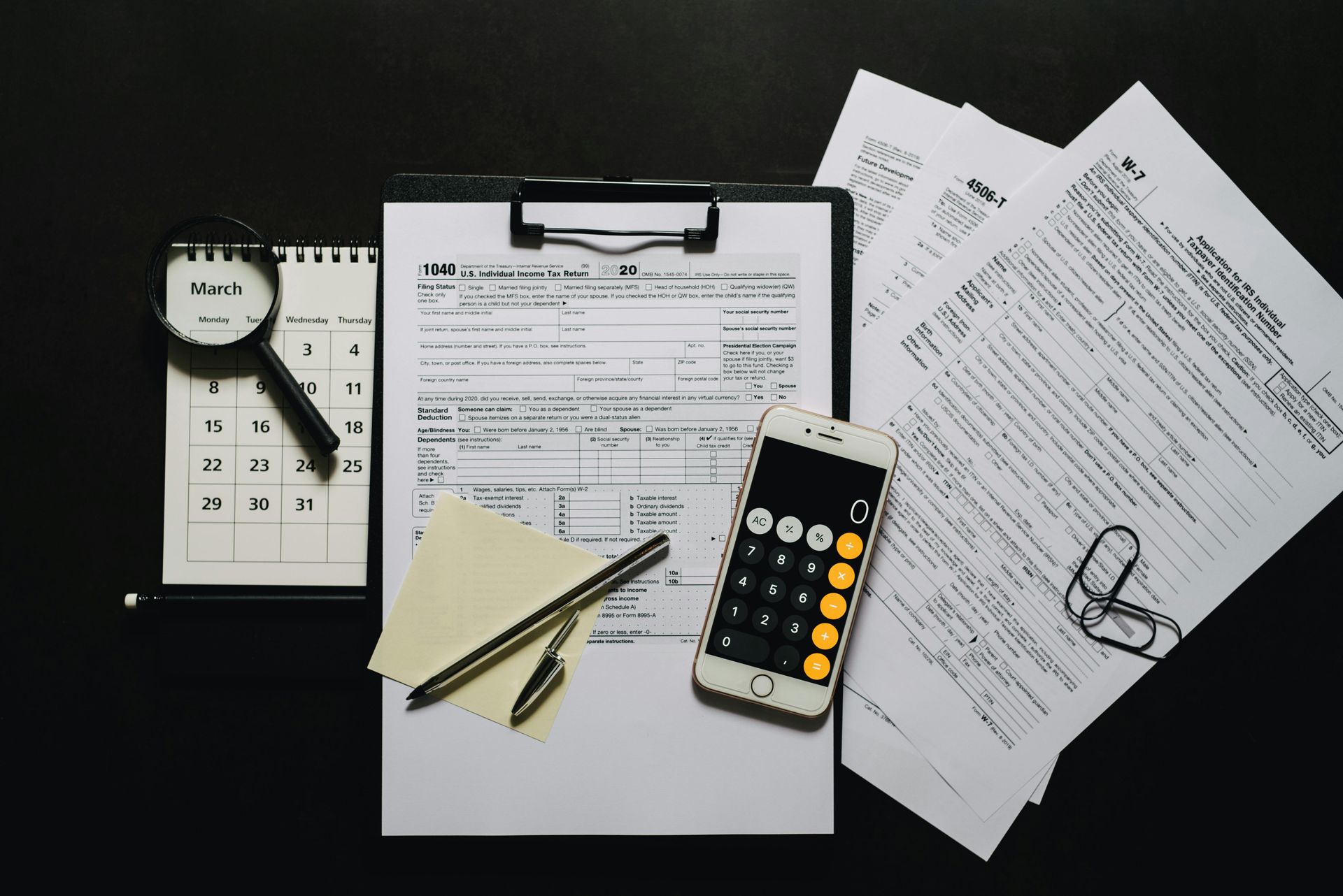 Tax documents, calculator, phone, and magnifying glass on a dark surface, indicating tax preparation.