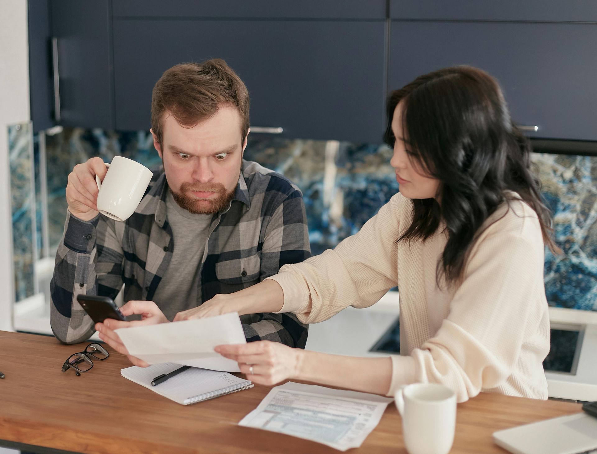 Man and woman reviewing bills at a counter; man looks concerned, holding a coffee cup.