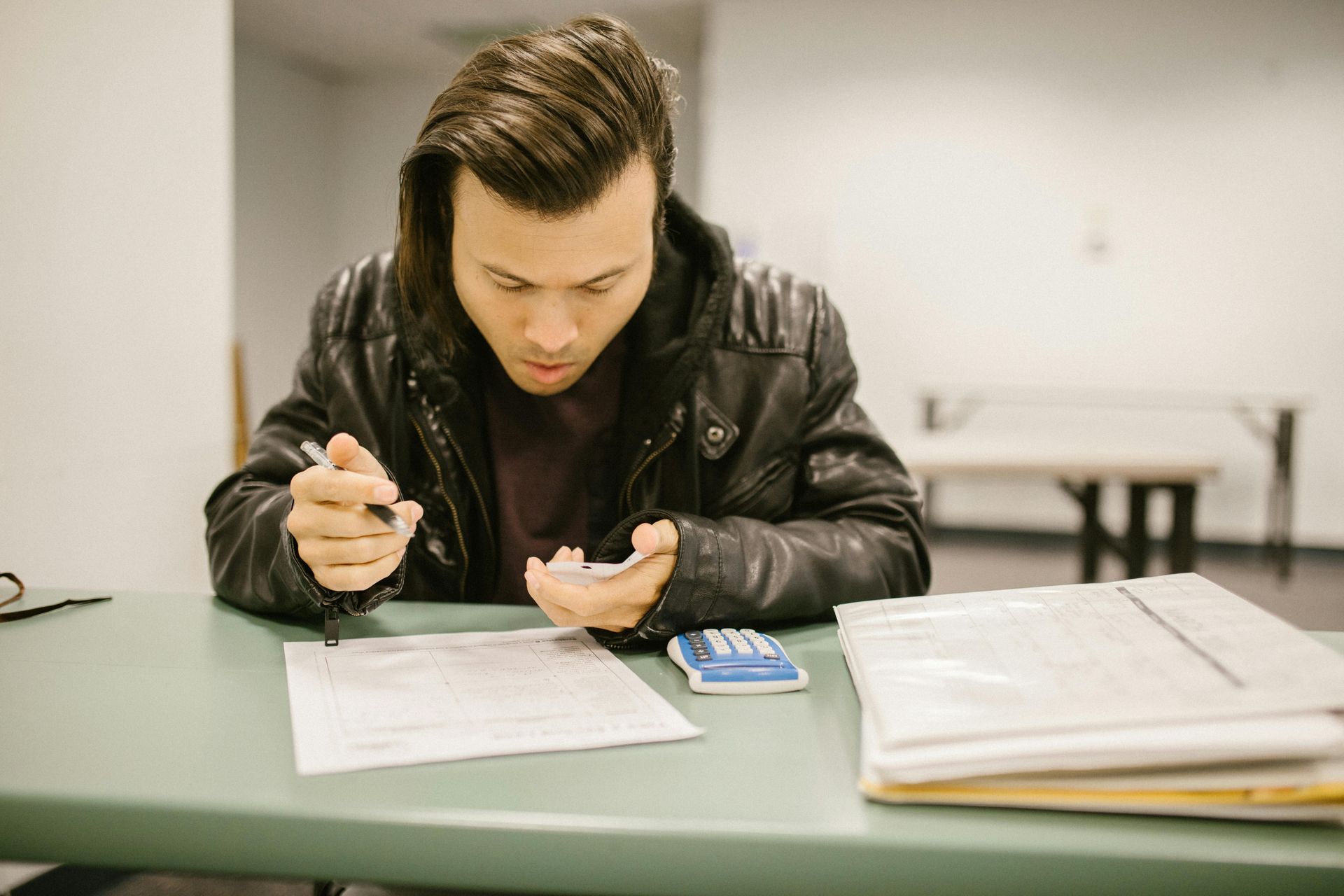 A person in a black leather jacket sits at a table, focused on writing on a document with a calculator nearby.