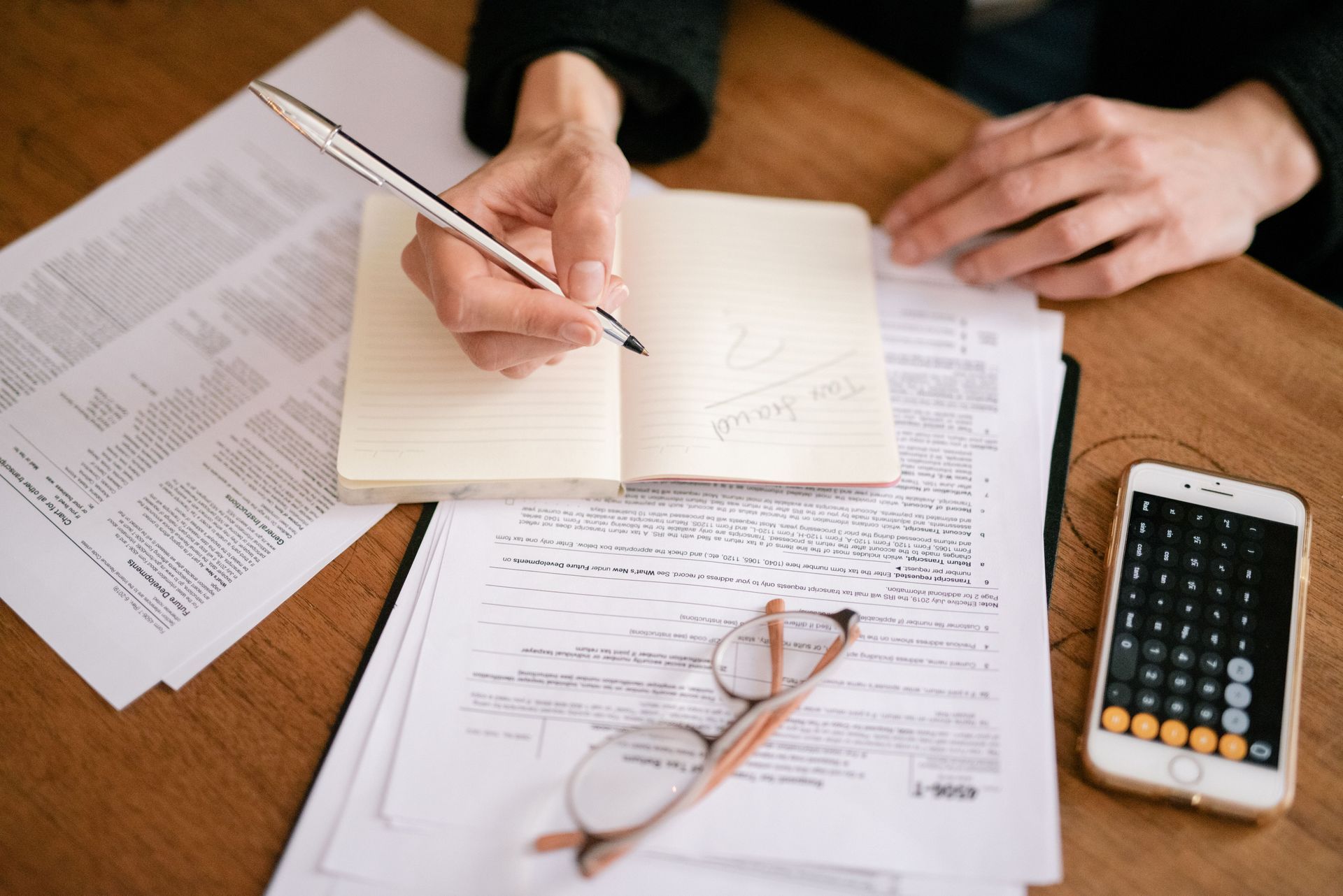 Hands writing in an open notebook amid papers, glasses, and a calculator on a desk