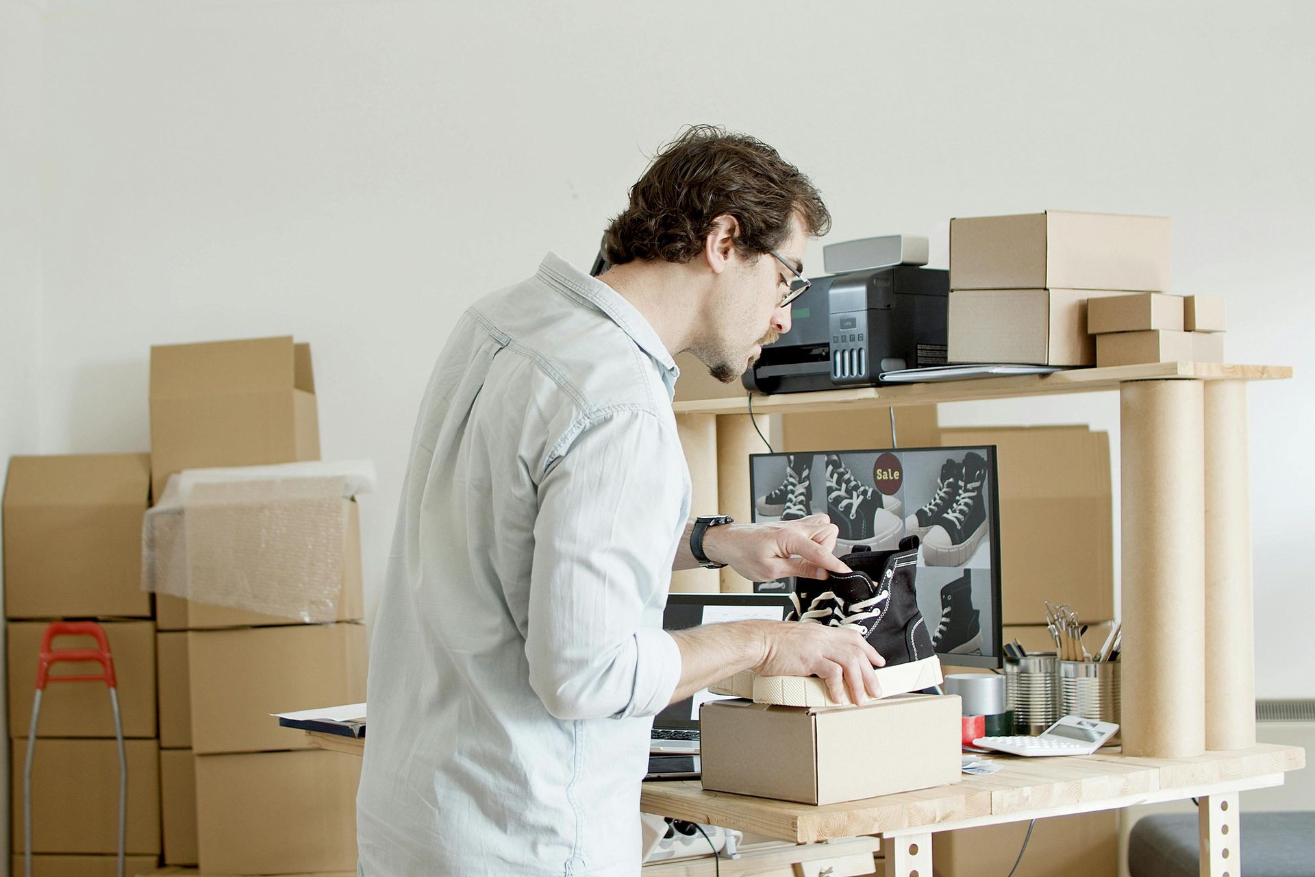 A person wearing a light shirt stands at a wooden workstation, packing a cardboard box among stacks of shipping supplies.