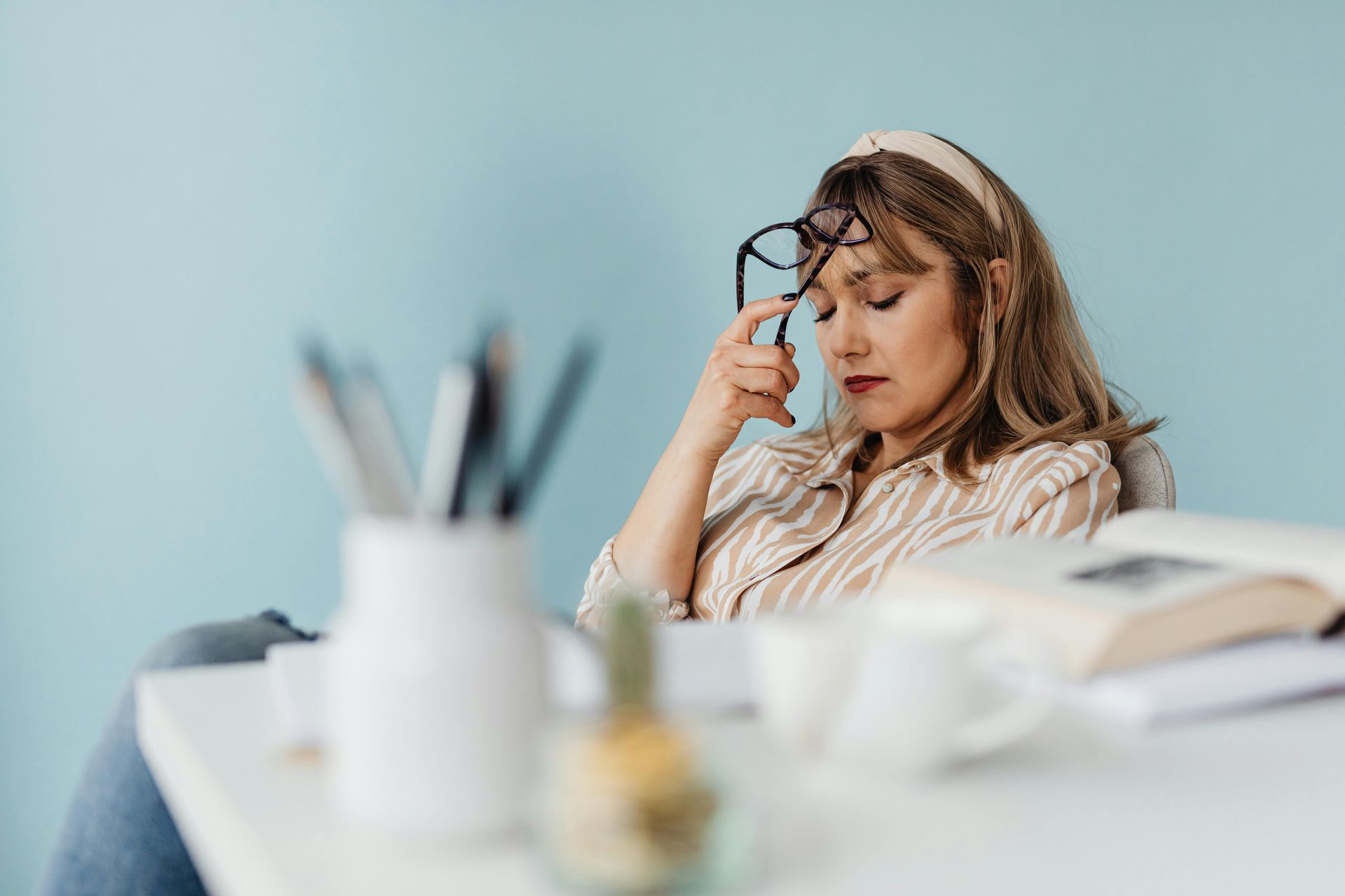 Woman resting head, holding glasses, at a desk with books and pens; light blue wall.