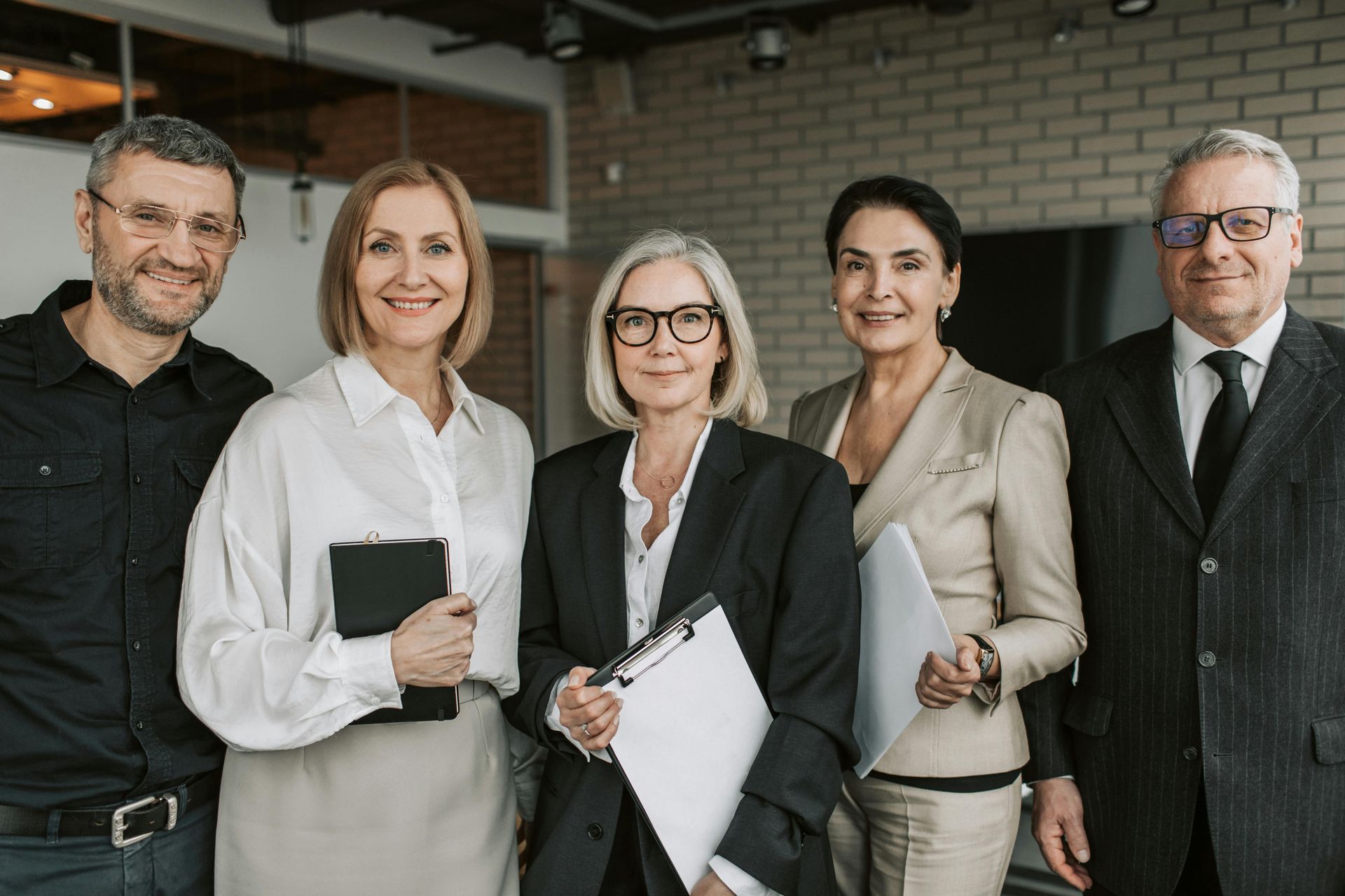 Five professionals smiling, in an office setting. Three women and two men wearing business attire.