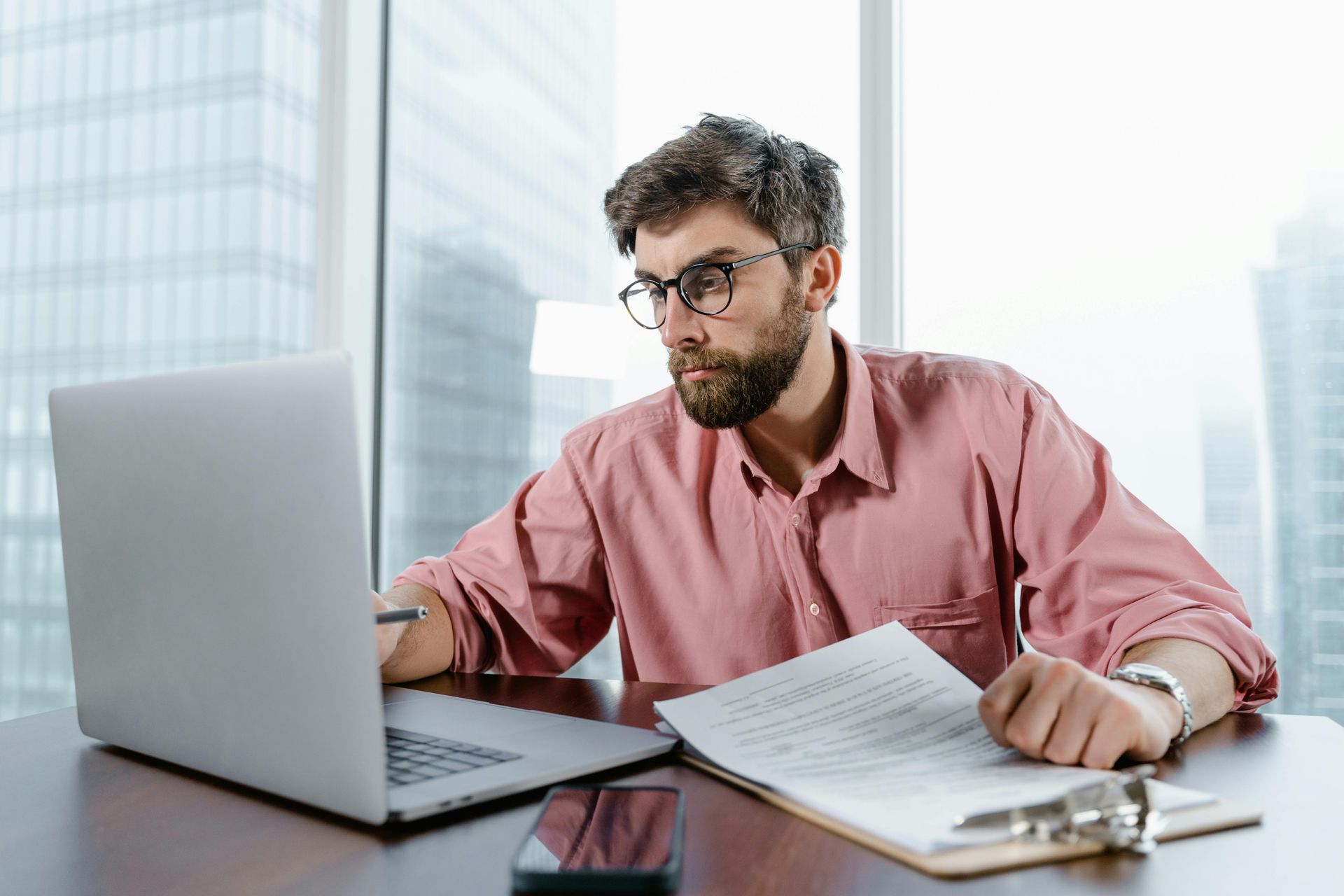Man with glasses, pink shirt, working on laptop, reviewing paperwork, city backdrop.