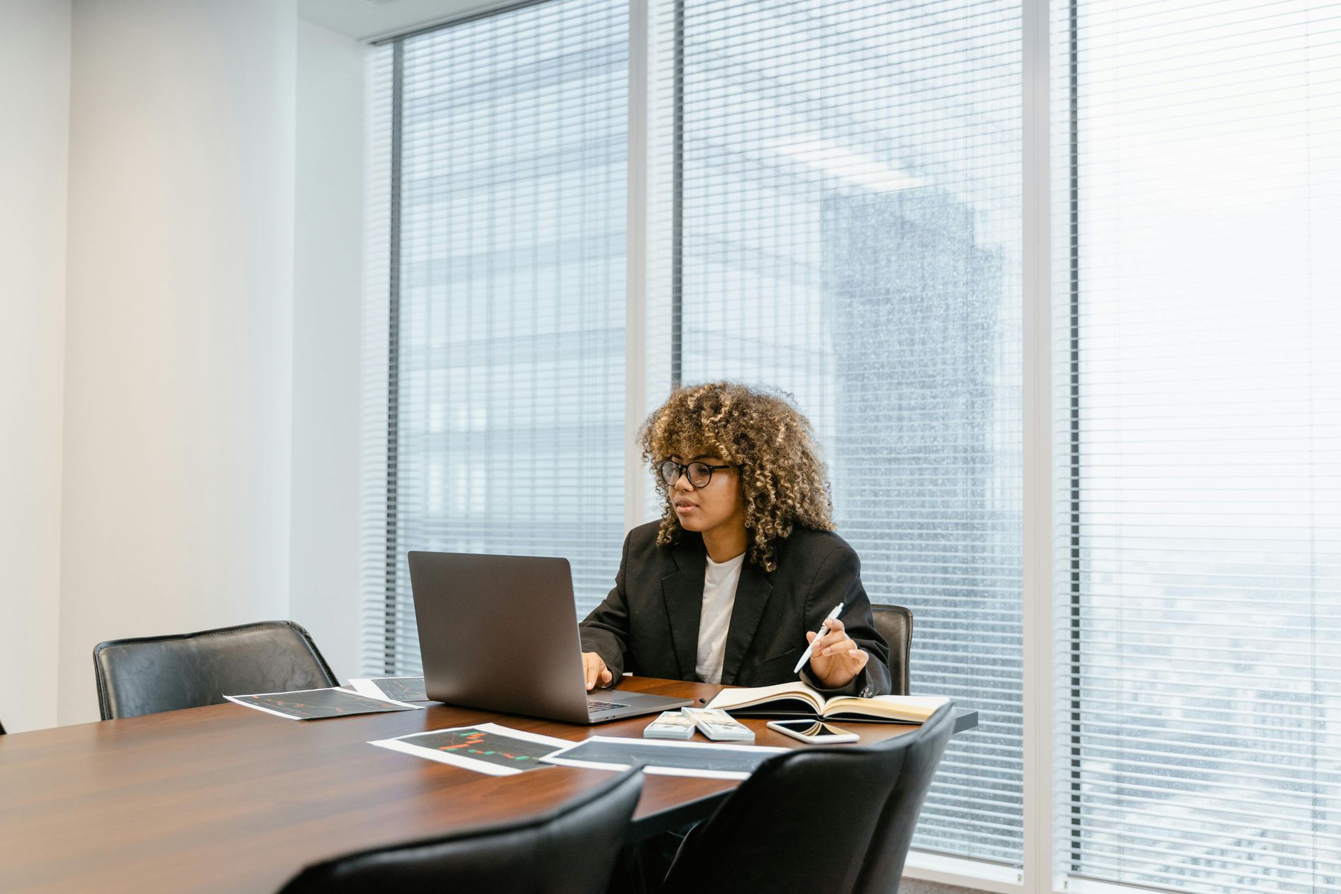 A person with curly hair and glasses works on a laptop in a modern office conference room with large window blinds.