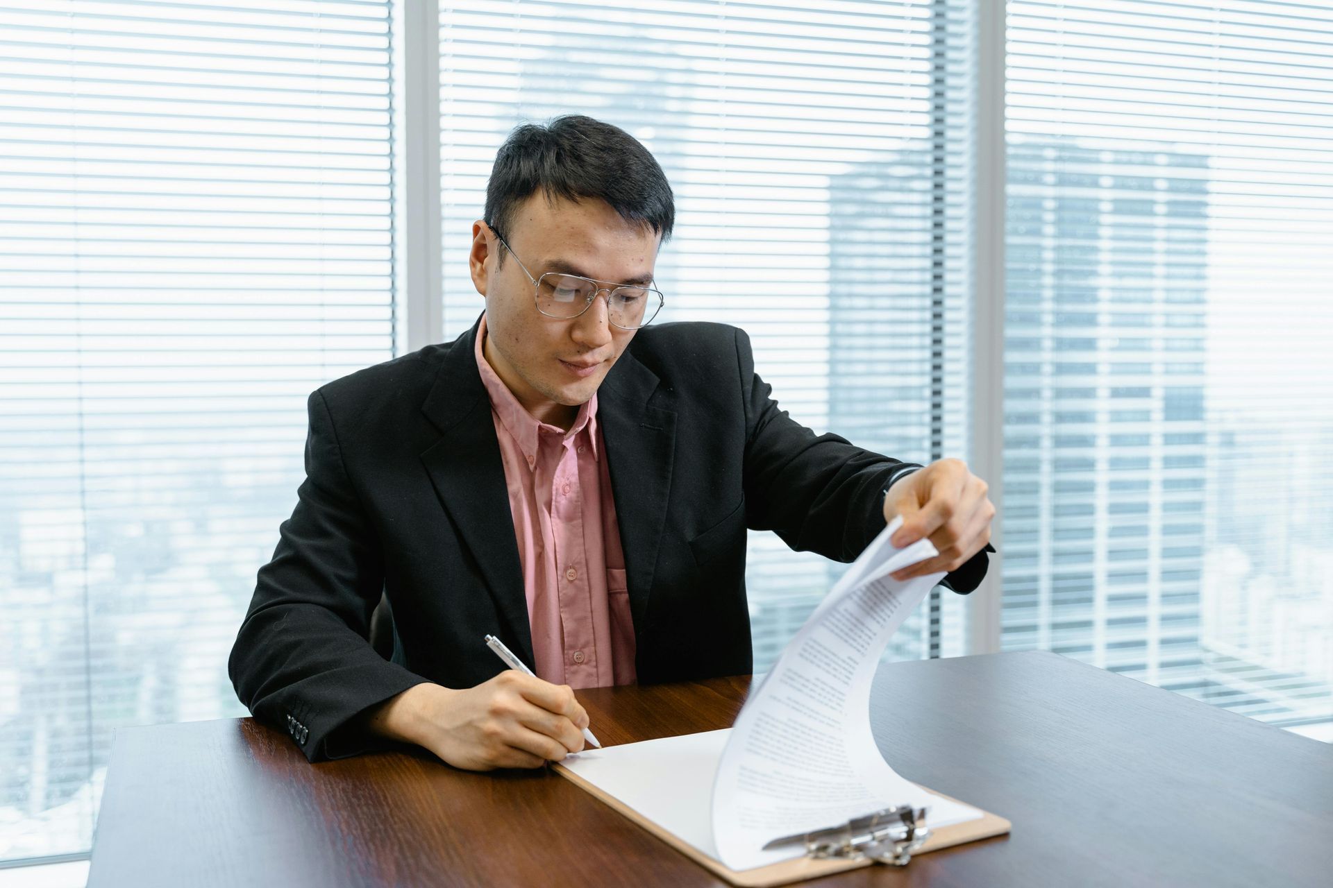 Man in blazer, signing papers on a clipboard at a desk in an office.