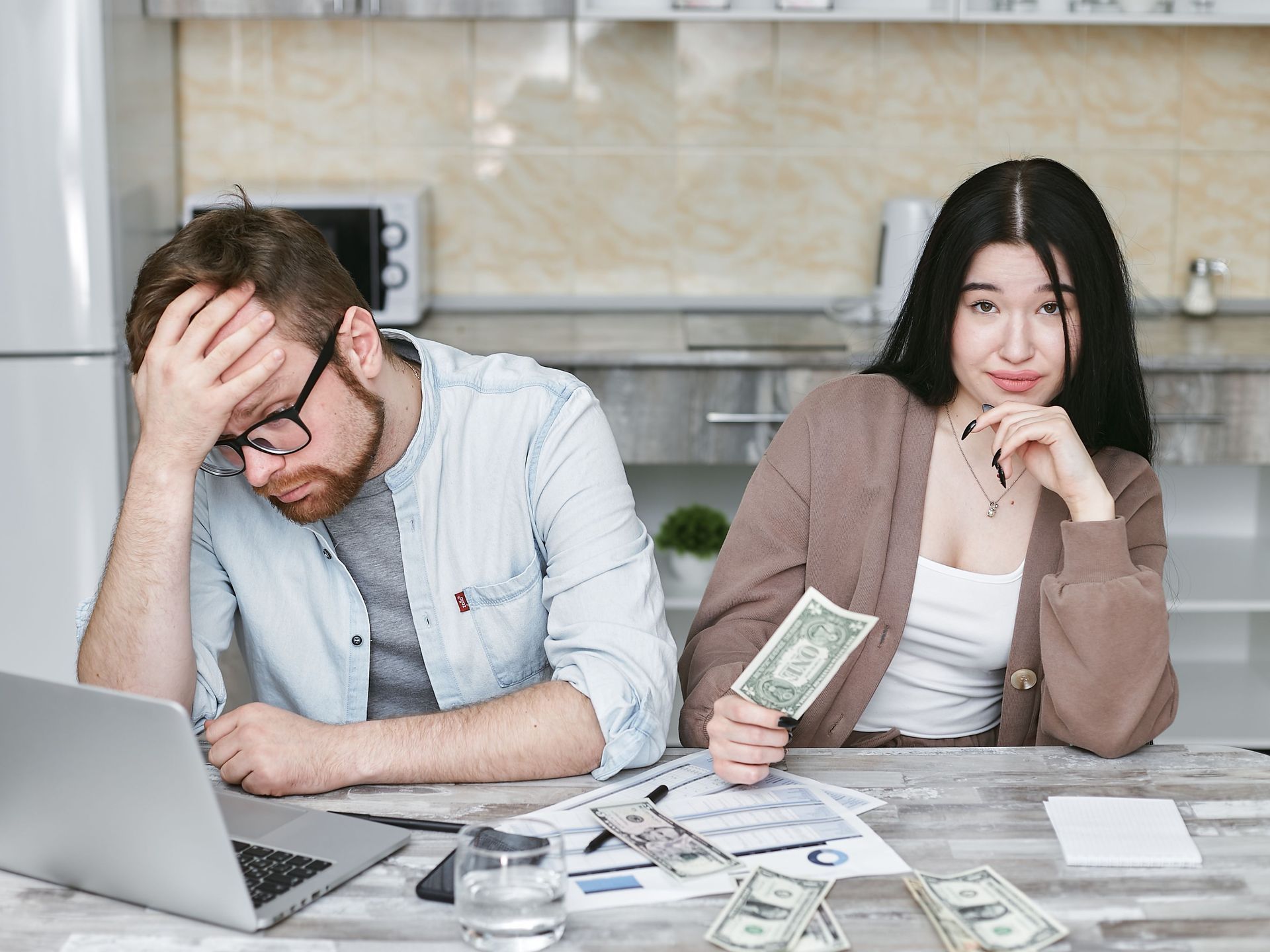 Stressed person at a laptop while a woman holds cash and looks on at a kitchen table with bills and papers