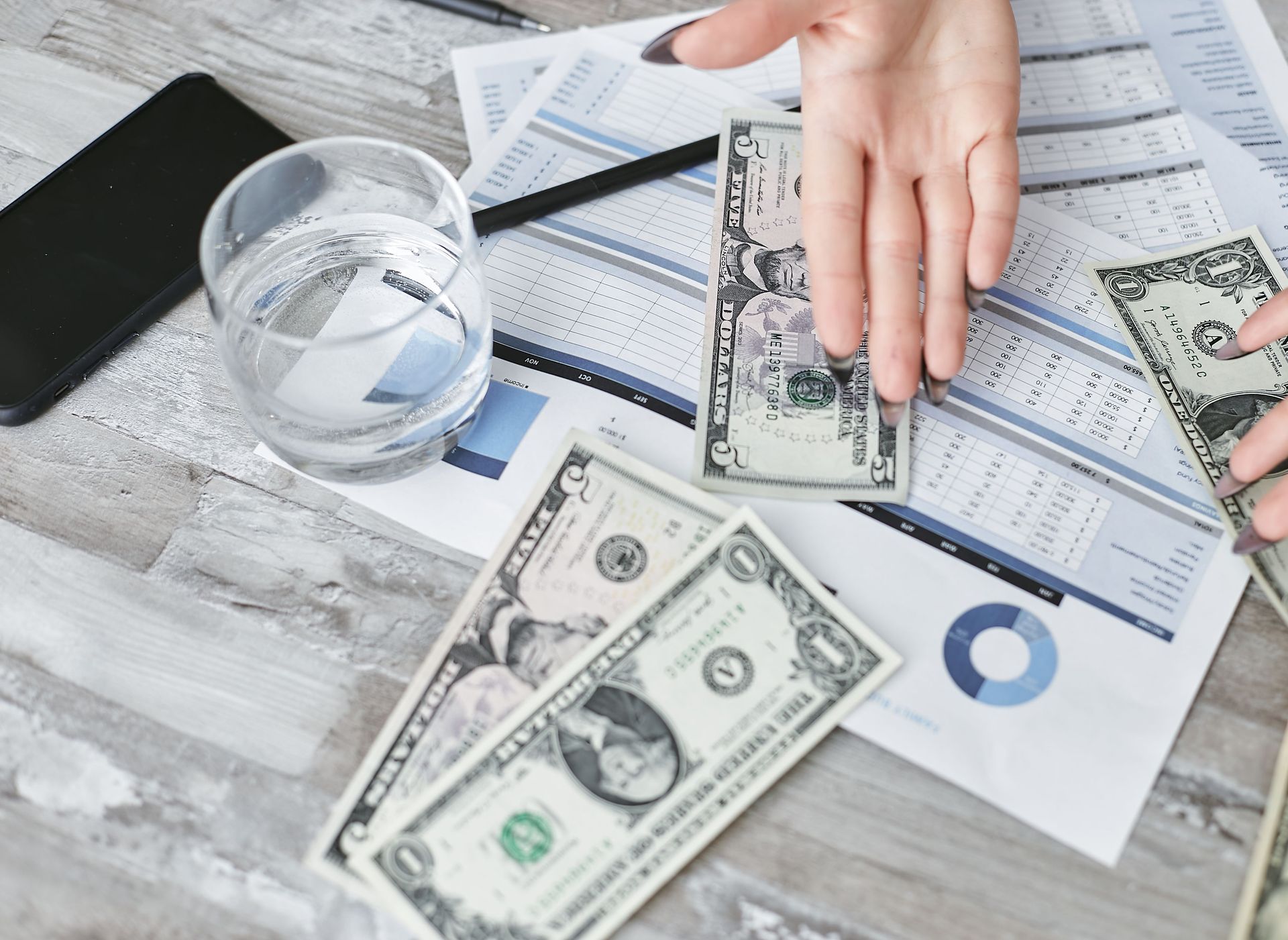 Hand holding dollar bills over a desk with papers, calculator, water glass, and scattered cash