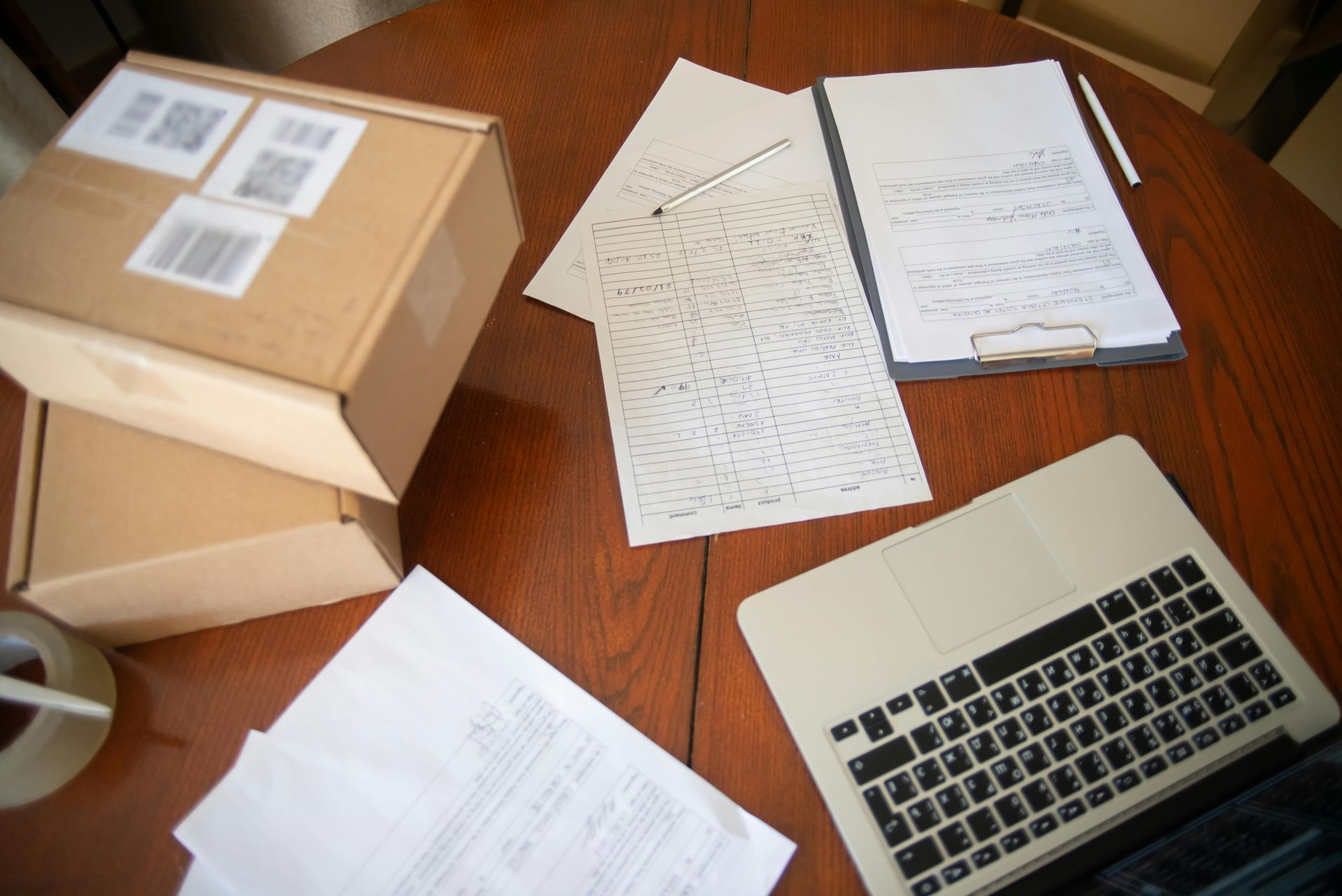 Two stacked shipping boxes, handwritten papers, and a laptop arranged on a circular wooden table.