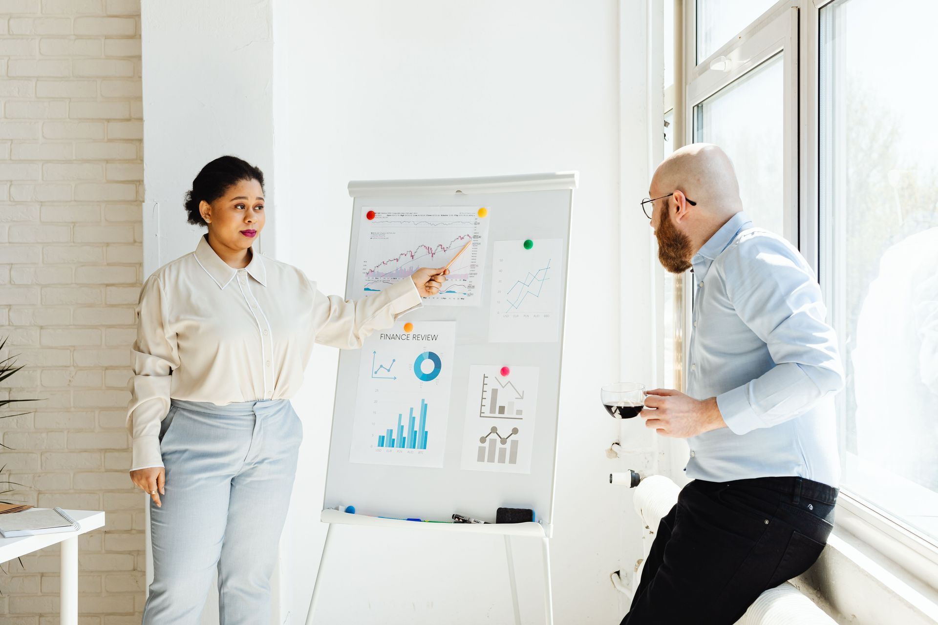 Two coworkers discuss charts on a whiteboard in a bright office, one pointing while the other listens.