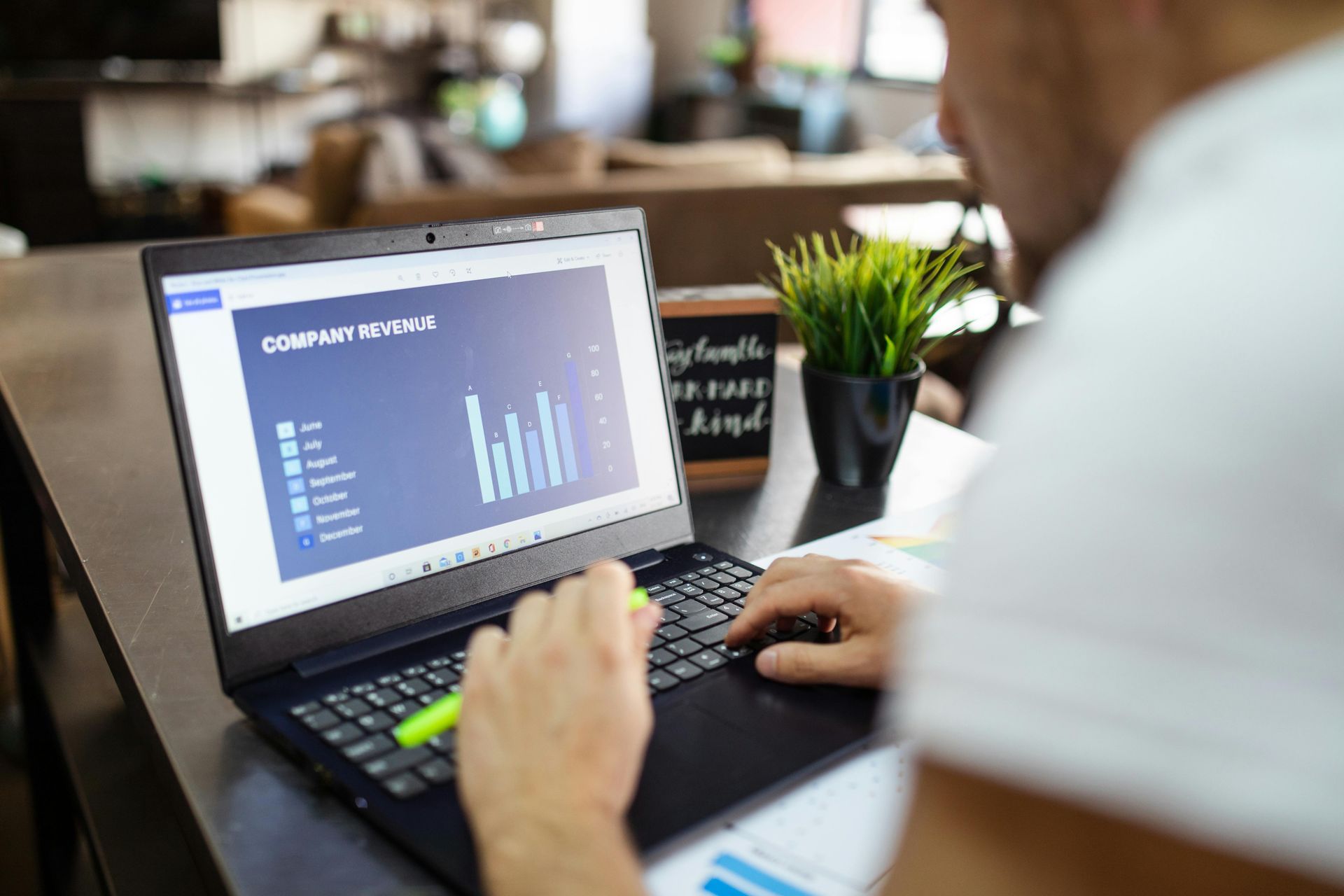 Person using a laptop, reviewing a bar graph with a highlighter in hand, at a table near a plant.