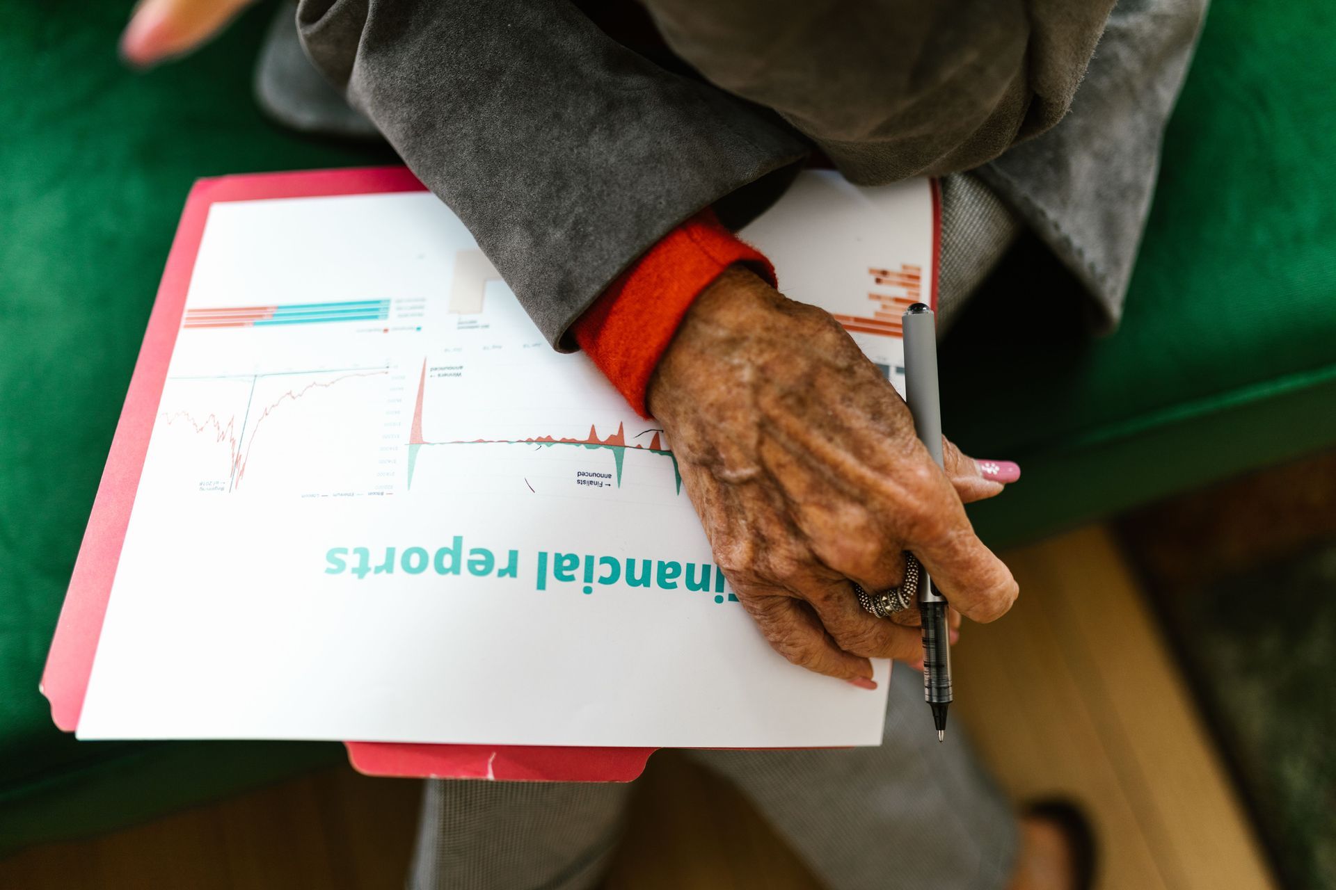 Hand holding a pen over a binder with financial reports on a green couch