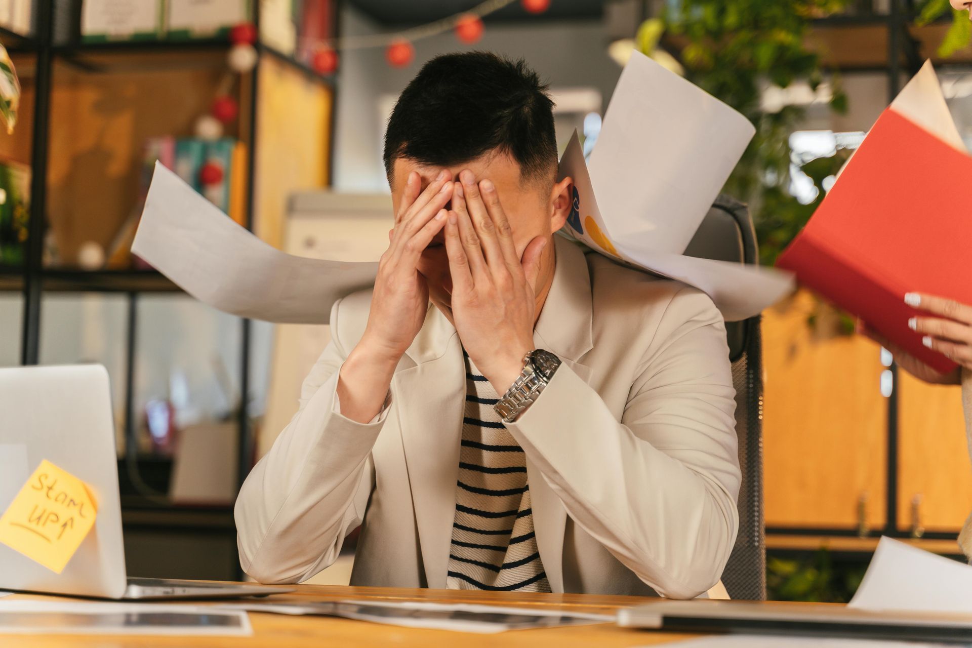 Stressed person at desk with hands covering face amid flying papers in an office