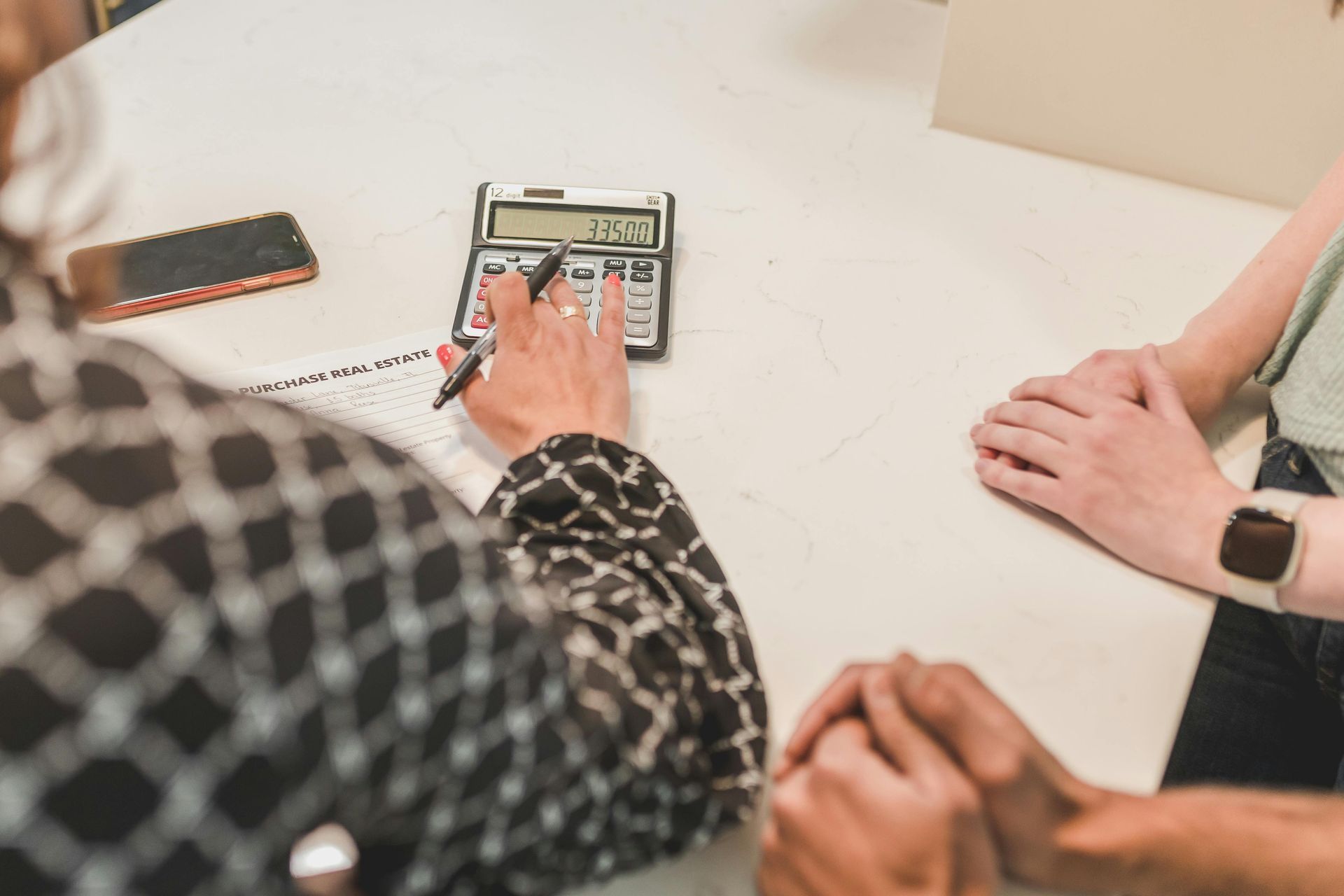 Person using a calculator, assisting another person at a table, with paperwork and a smartphone visible.