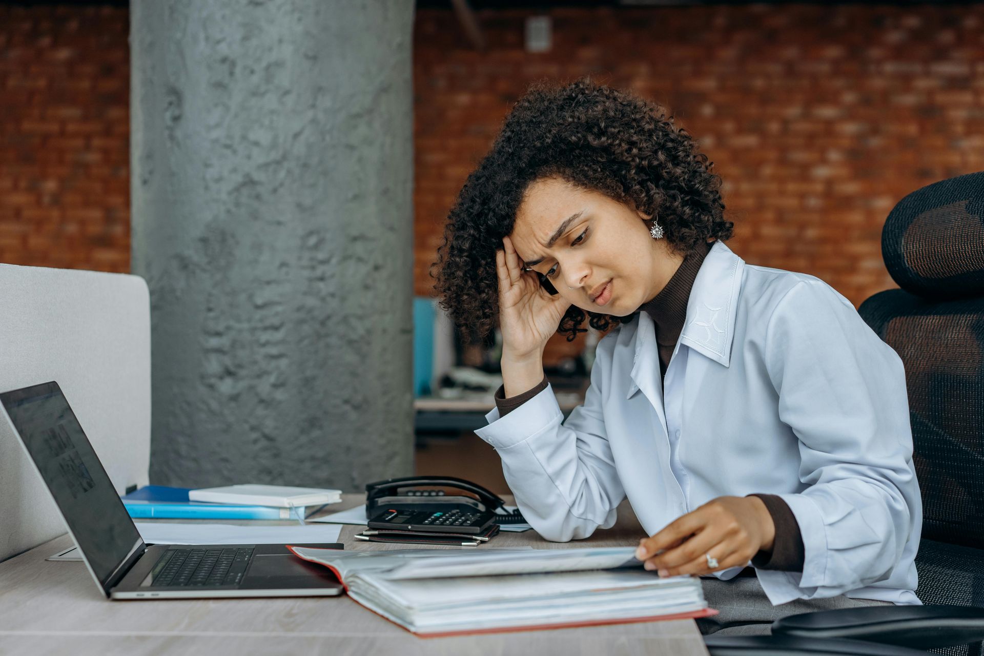 A person sitting at a desk with a laptop and open file, appearing stressed while holding their head in their hand.